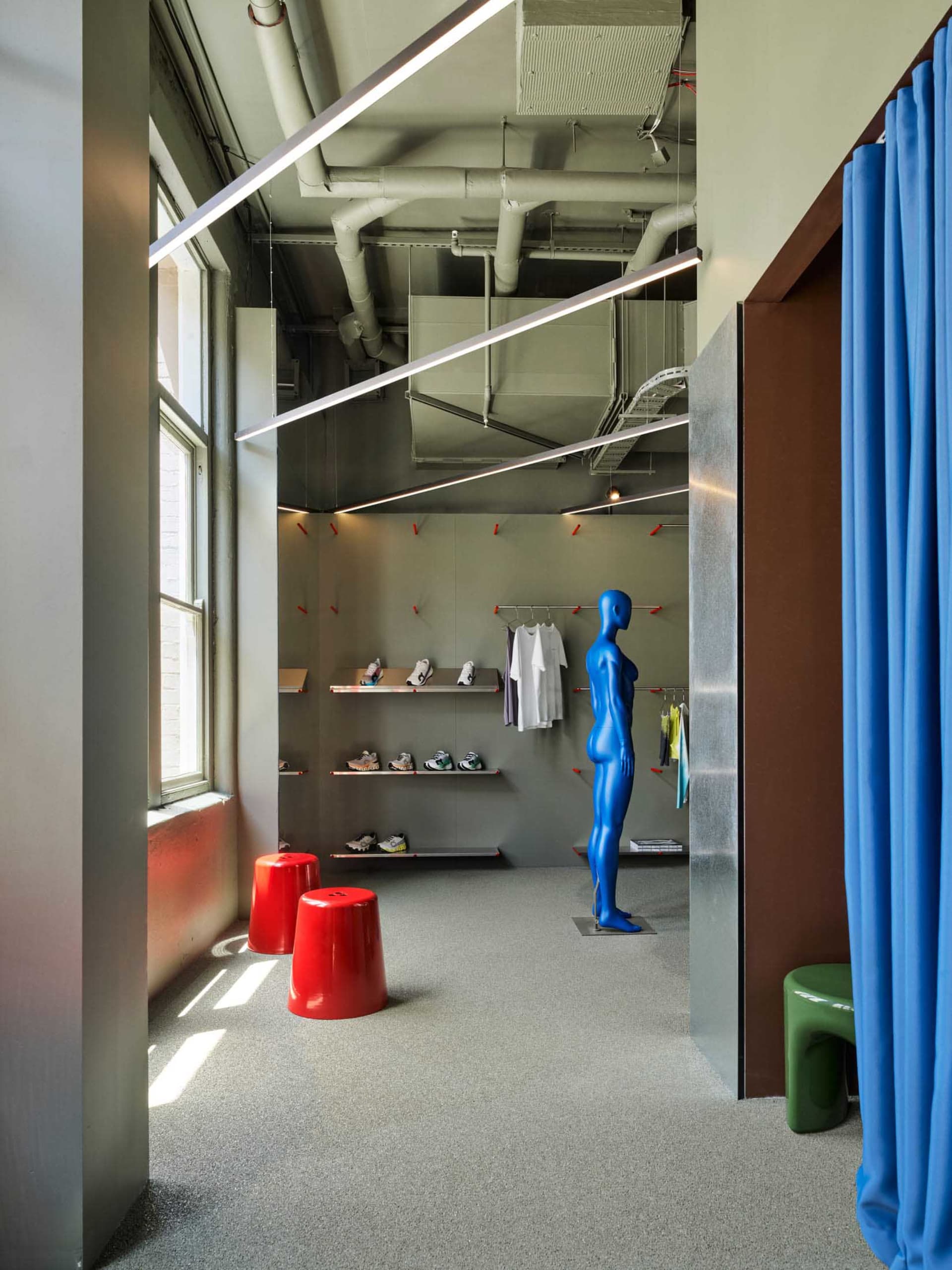Interior view of a modern industrial-style sneaker store featuring a vibrant blue mannequin, floating shelves with sneakers, clothing racks, bright red stools, and exposed ceiling pipes.