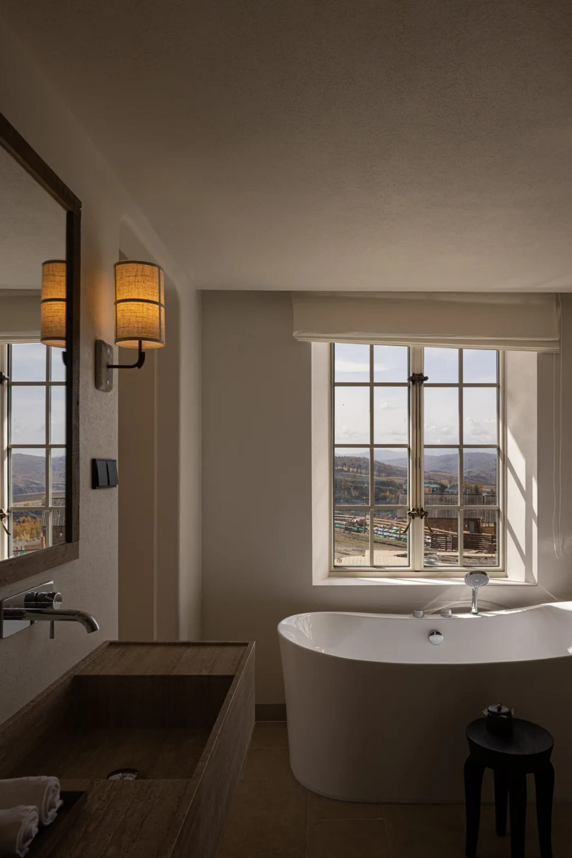 Modern bathroom interior featuring a white freestanding bathtub, wooden vanity with vessel sink, wall sconces, and large window overlooking mountains.