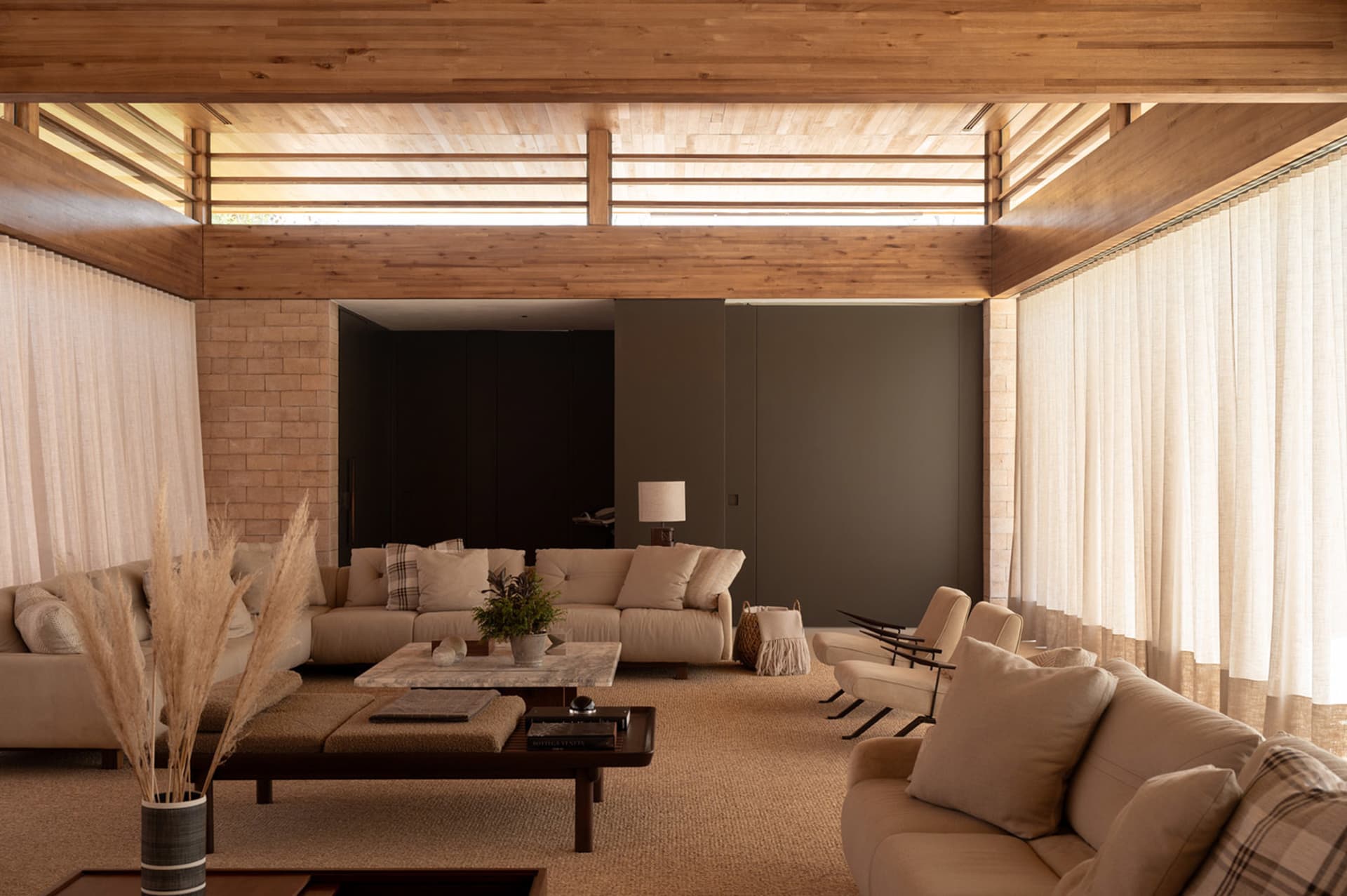 Modern organic living room featuring a high wood-paneled ceiling, exposed brick walls, neutral sectional sofas, marble coffee table, and abundant natural light from large windows with sheer curtains.