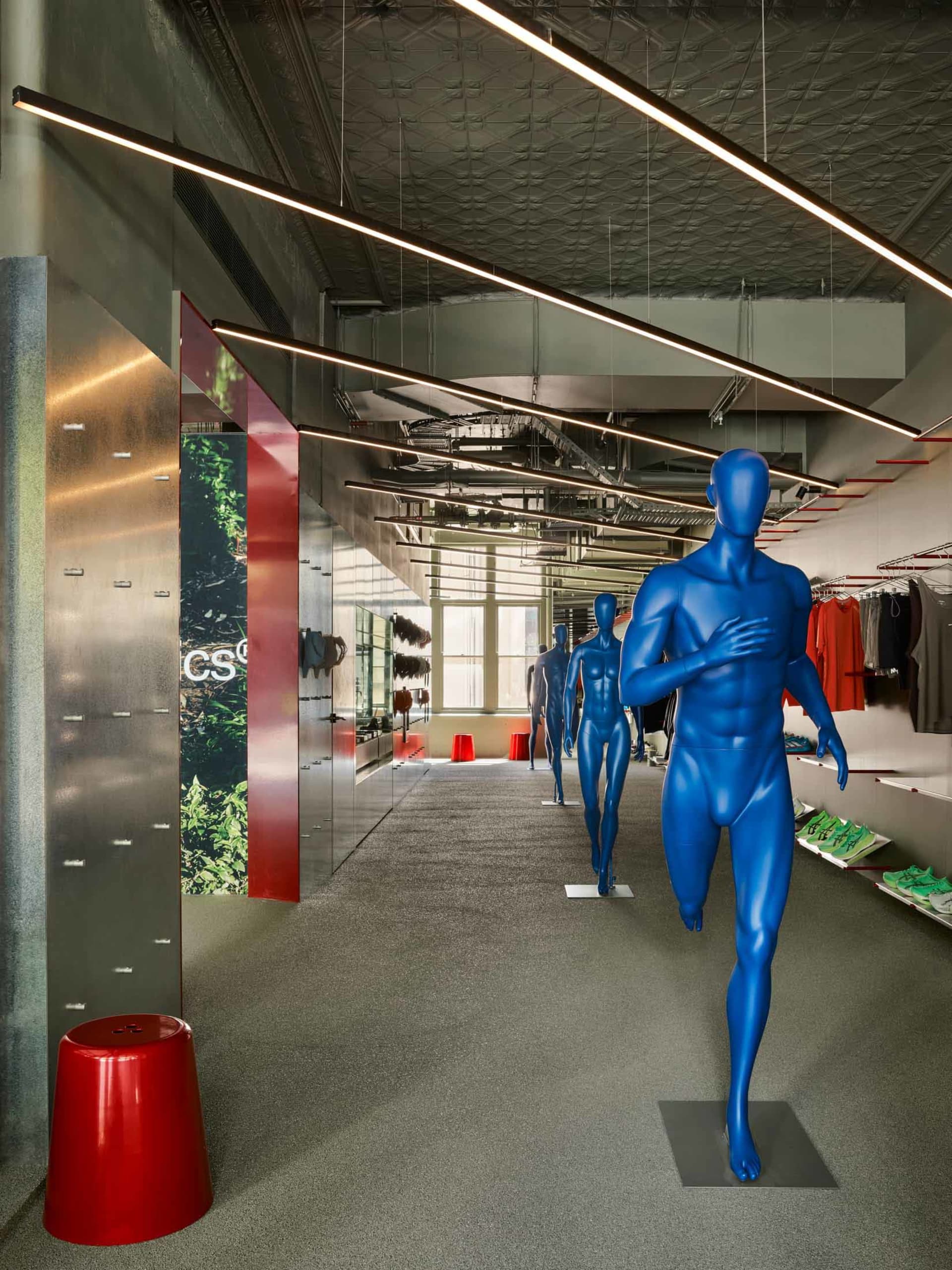 Interior view of a modern industrial-style retail store featuring multiple blue male mannequins, a bright red stool, linear LED lighting, and exposed ceiling infrastructure.