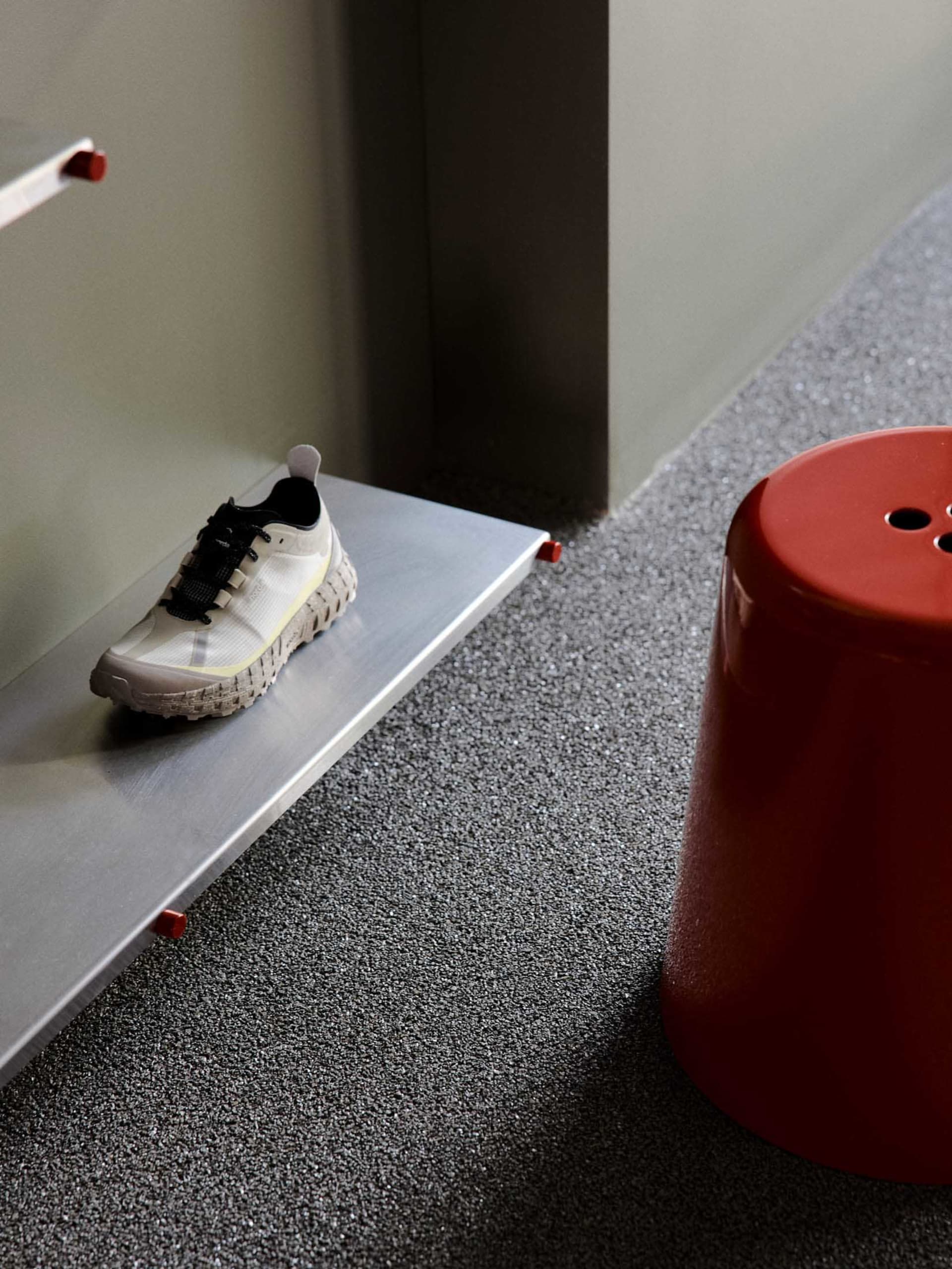 White and black athletic shoe resting on a stainless steel floating shelf above dark grey speckled carpet, with a bright red cylindrical stool beside it.