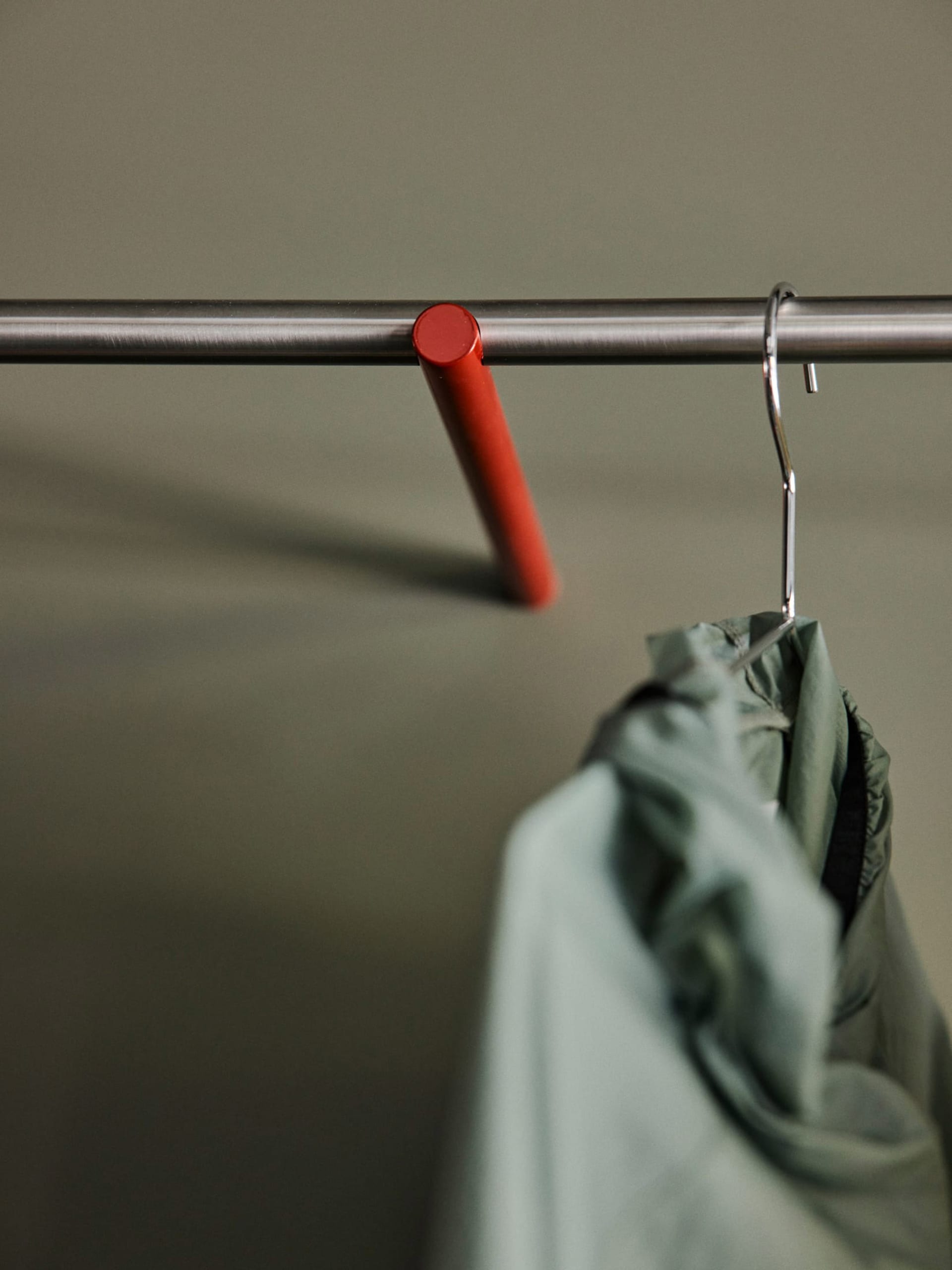 Close-up of a sage green jacket hanging on a chrome metal hanger from a red cylindrical hook attached to a brushed stainless steel clothing rack against a muted green wall.