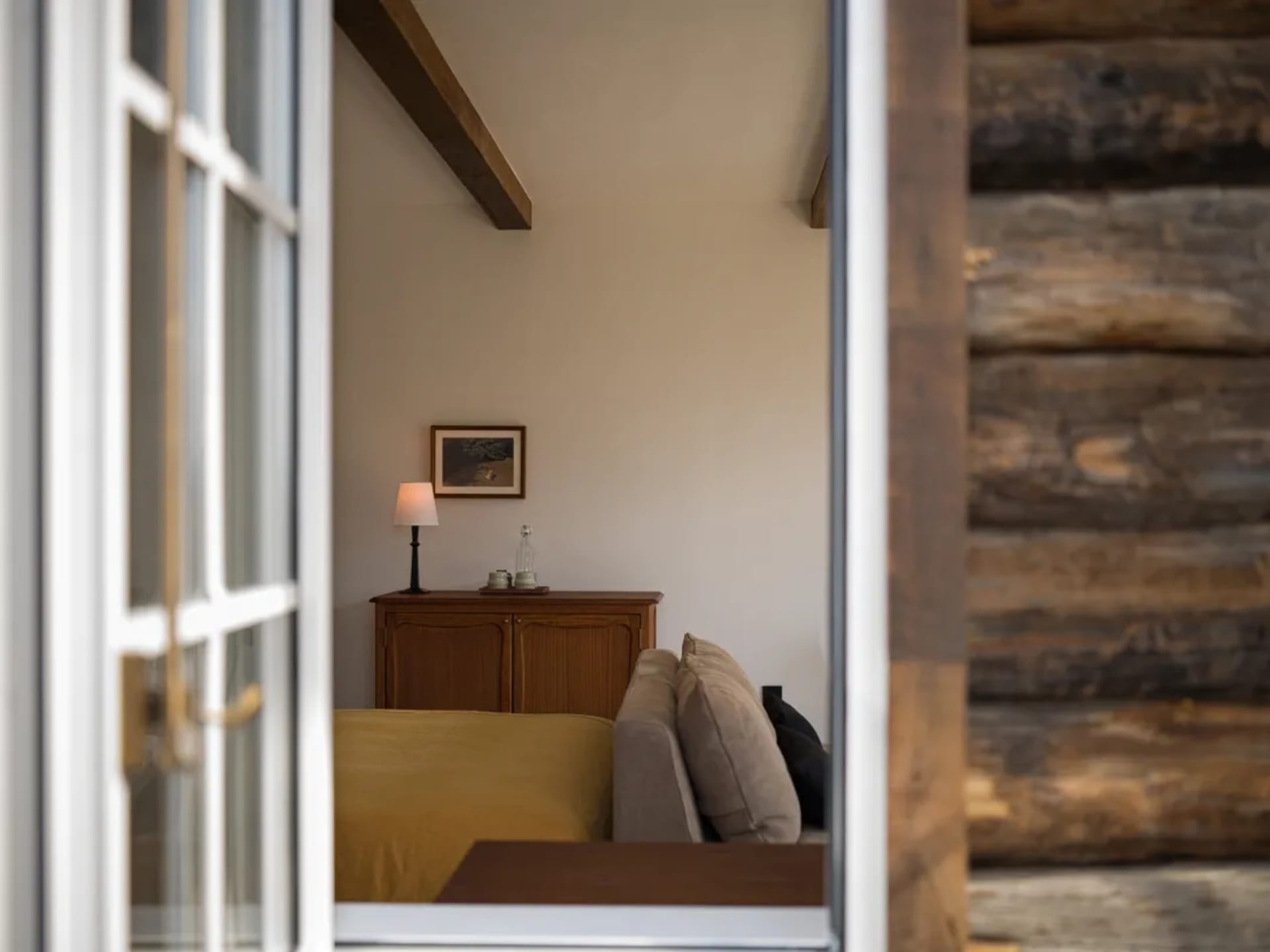 View through a white French door into a cozy cabin living room featuring a mustard-yellow sofa, beige linen sofa, reclaimed wood accent wall, exposed wooden beams, and a wooden sideboard with a lamp.