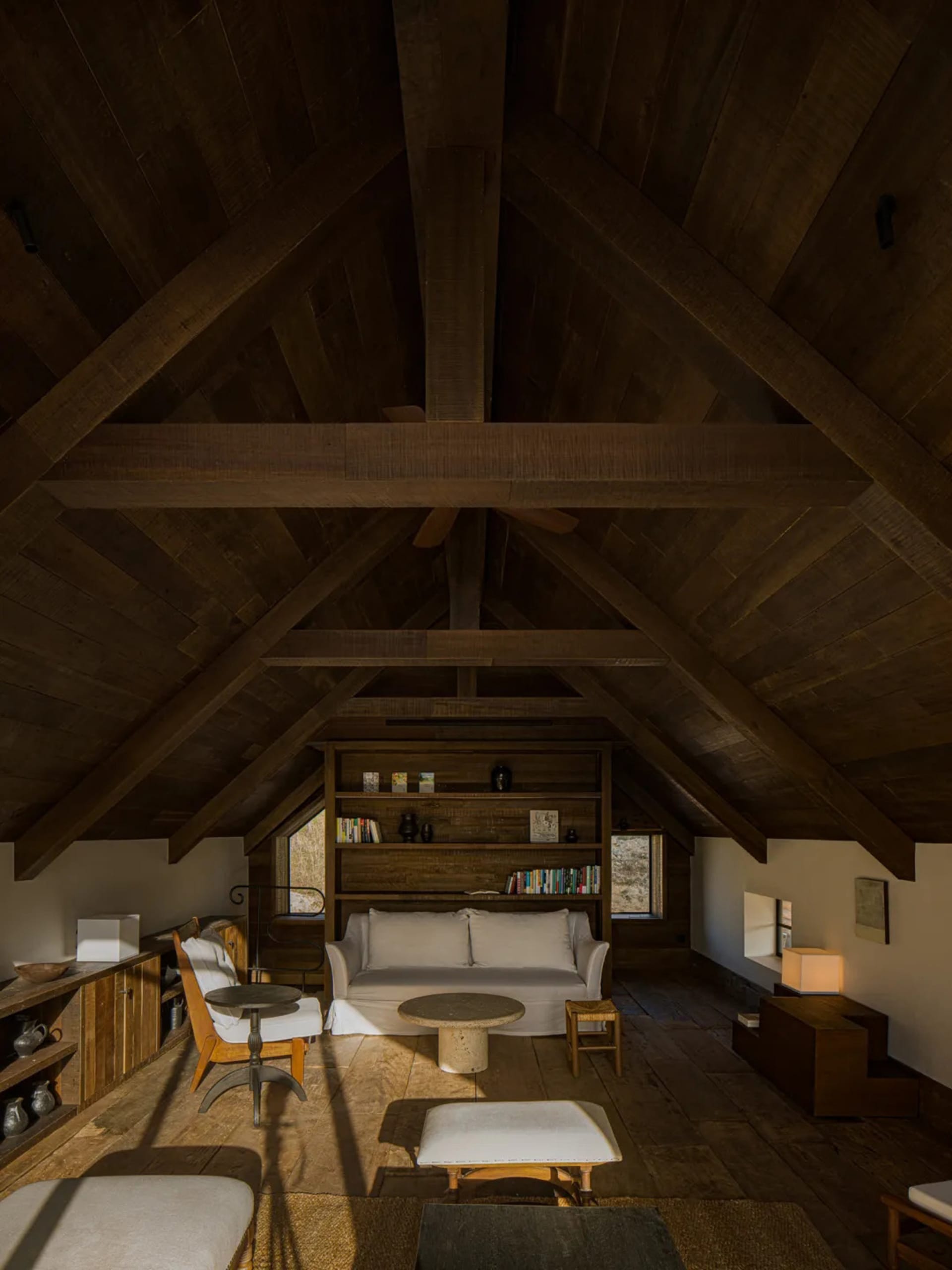 A spacious loft living room with high cathedral ceiling featuring massive reclaimed wood beams, a white sofa against a built-in wooden bookshelf, stone coffee table, and wooden flooring bathed in natural light.