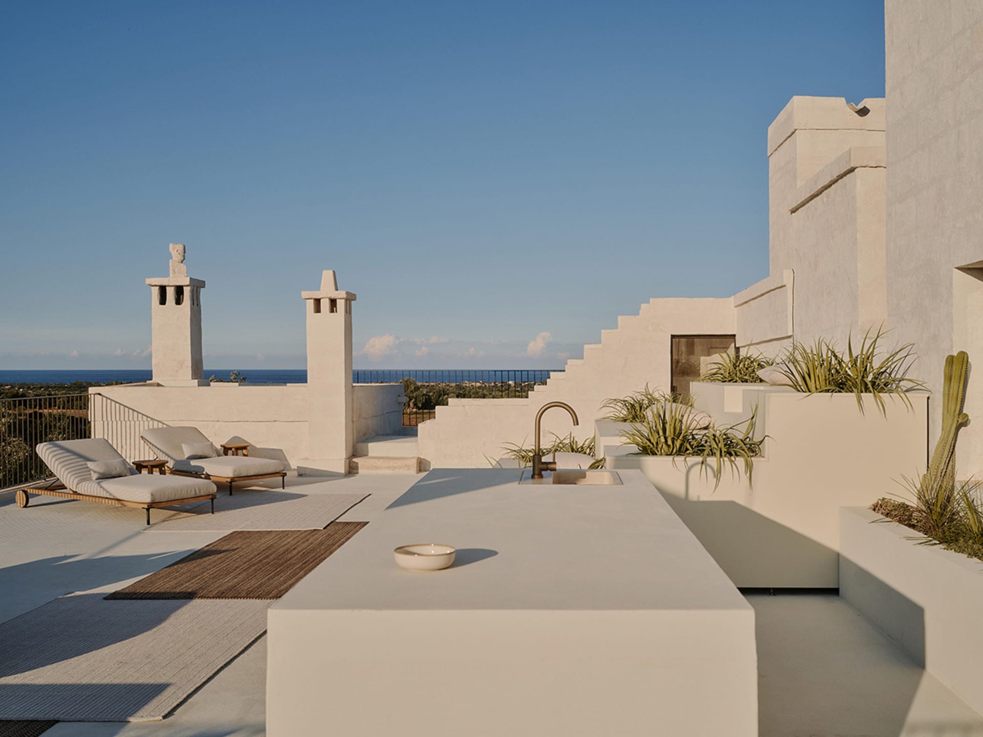 Modern minimalist rooftop terrace with white concrete outdoor kitchen island, built-in planters with cacti and grasses, wooden lounge chairs, and panoramic ocean view under a clear blue sky.