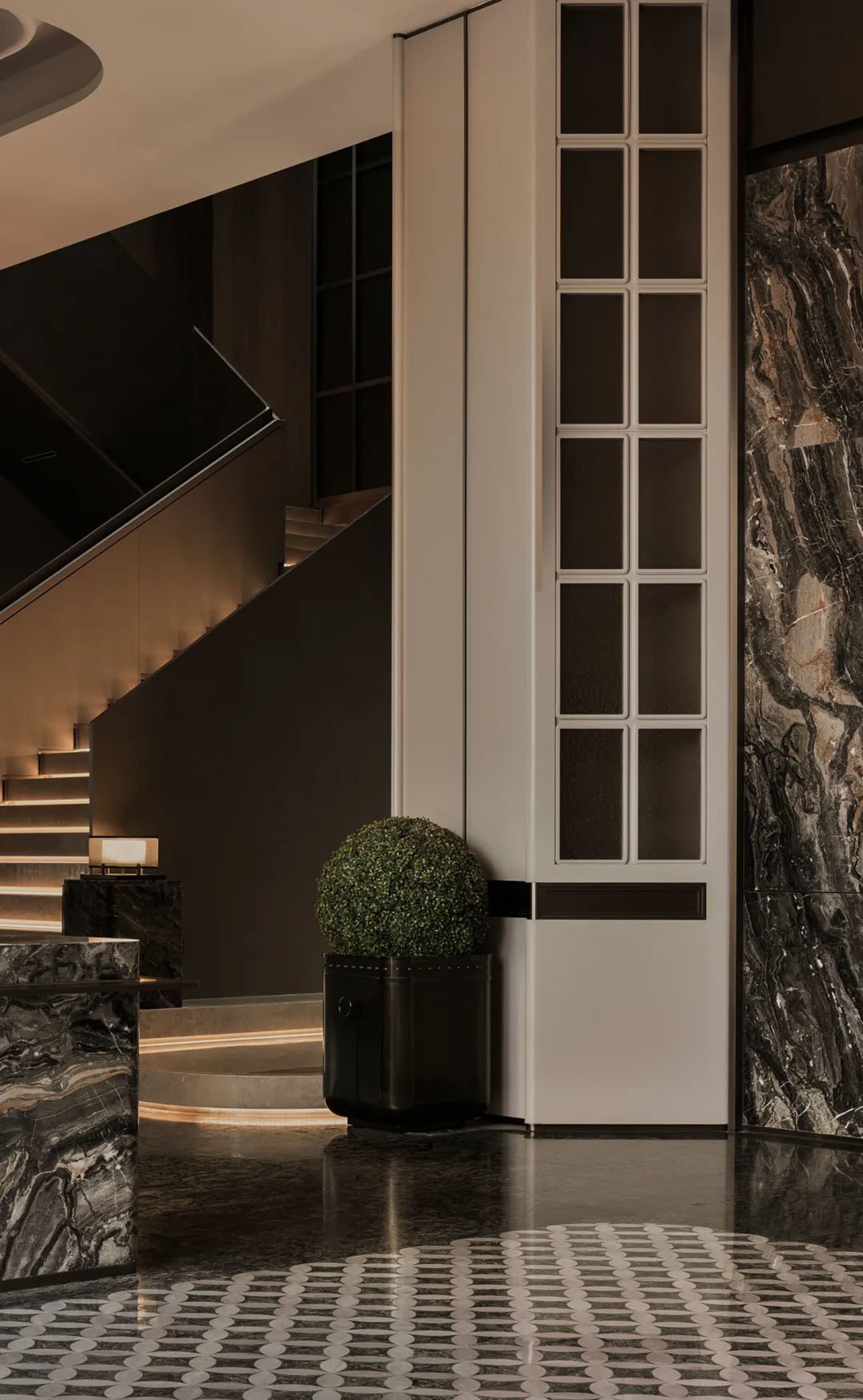 Modern luxury entrance hall featuring black marble floors and walls, a white paneled wall with grid windows, a spherical topiary in a black planter, and a floating staircase with warm LED step lighting.