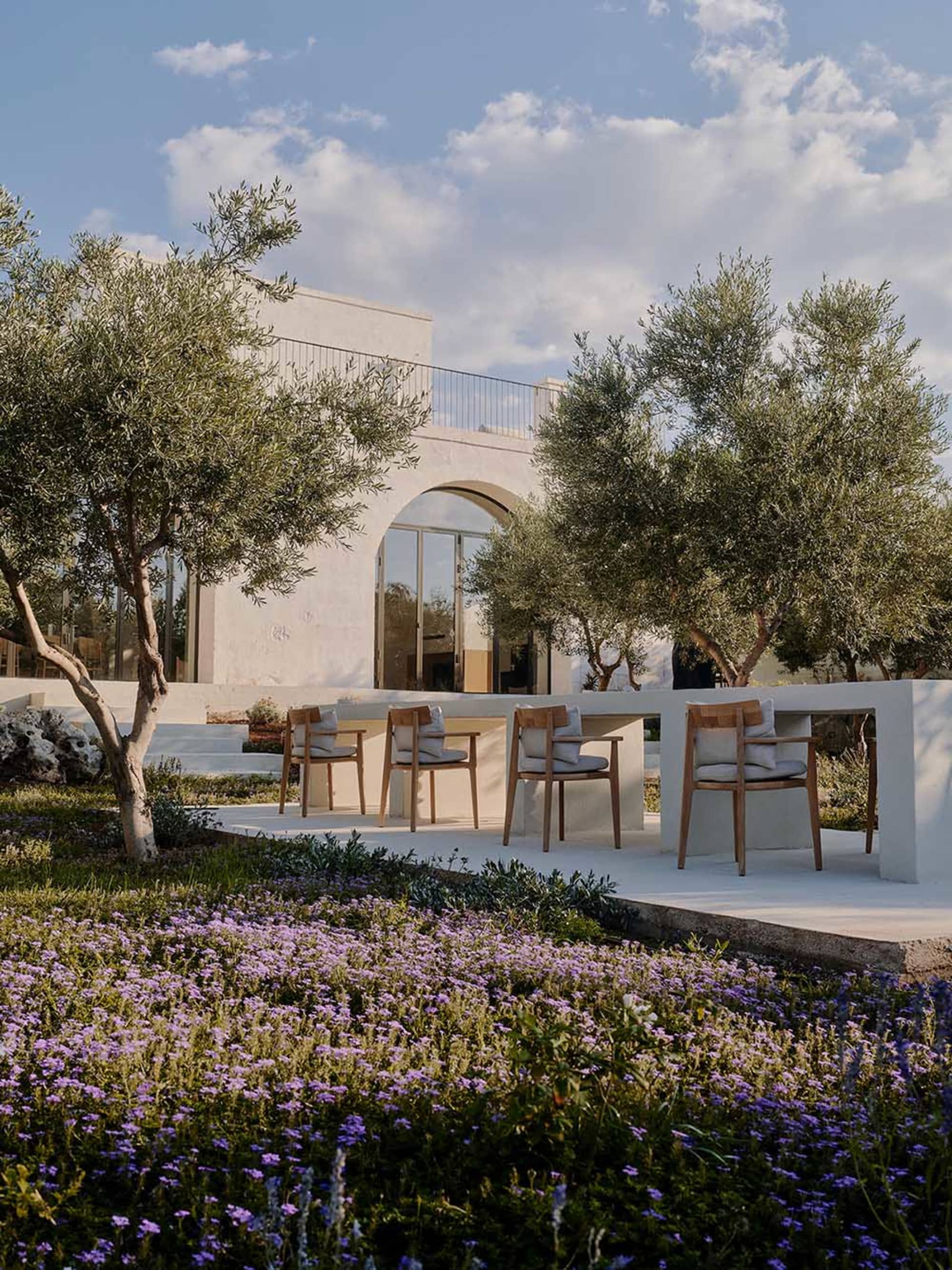 Modern Mediterranean villa terrace with wooden dining chairs, white limestone walls, olive trees, and a foreground of purple wildflowers under a bright blue sky.