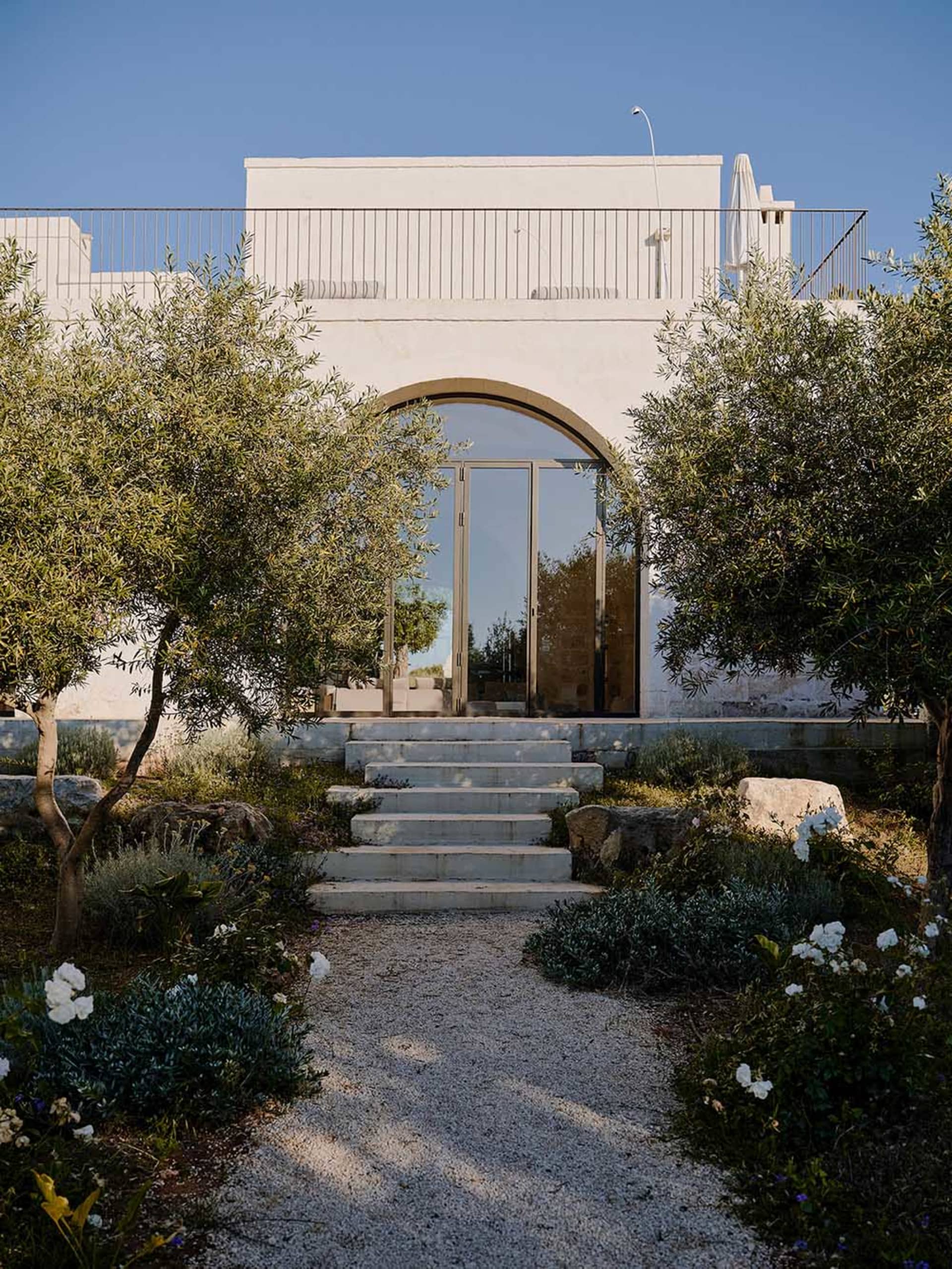 Front view of a modern white stucco villa with a large arched glass door, accessed by wide stone steps and surrounded by olive trees and Mediterranean garden plants under a clear blue sky.