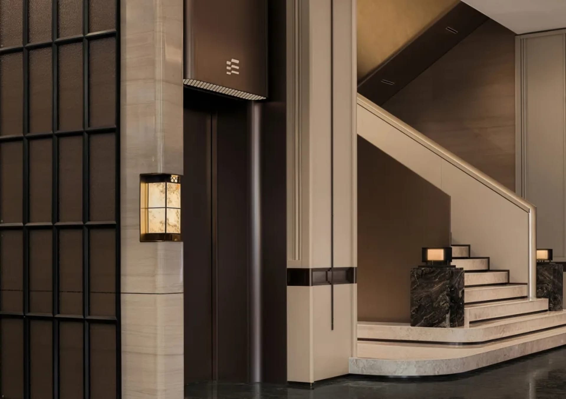 Modern luxury building lobby with beige marble walls, dark marble grand staircase, brass handrail, and warm alabaster wall sconces next to an elevator.
