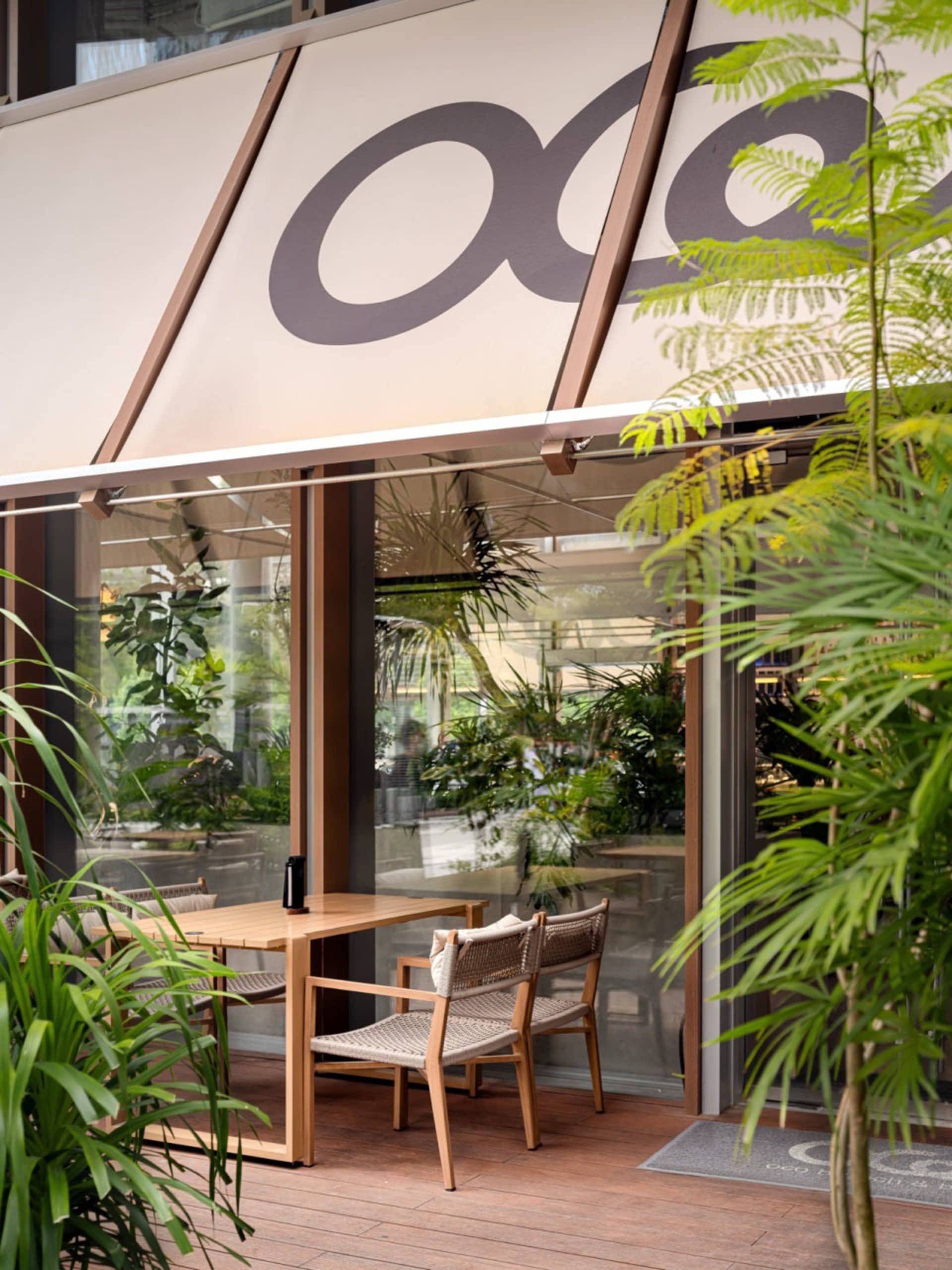 Modern outdoor cafe patio featuring a teak wood dining table, woven rattan chairs, lush tropical plants, and a large retractable fabric awning with logo.