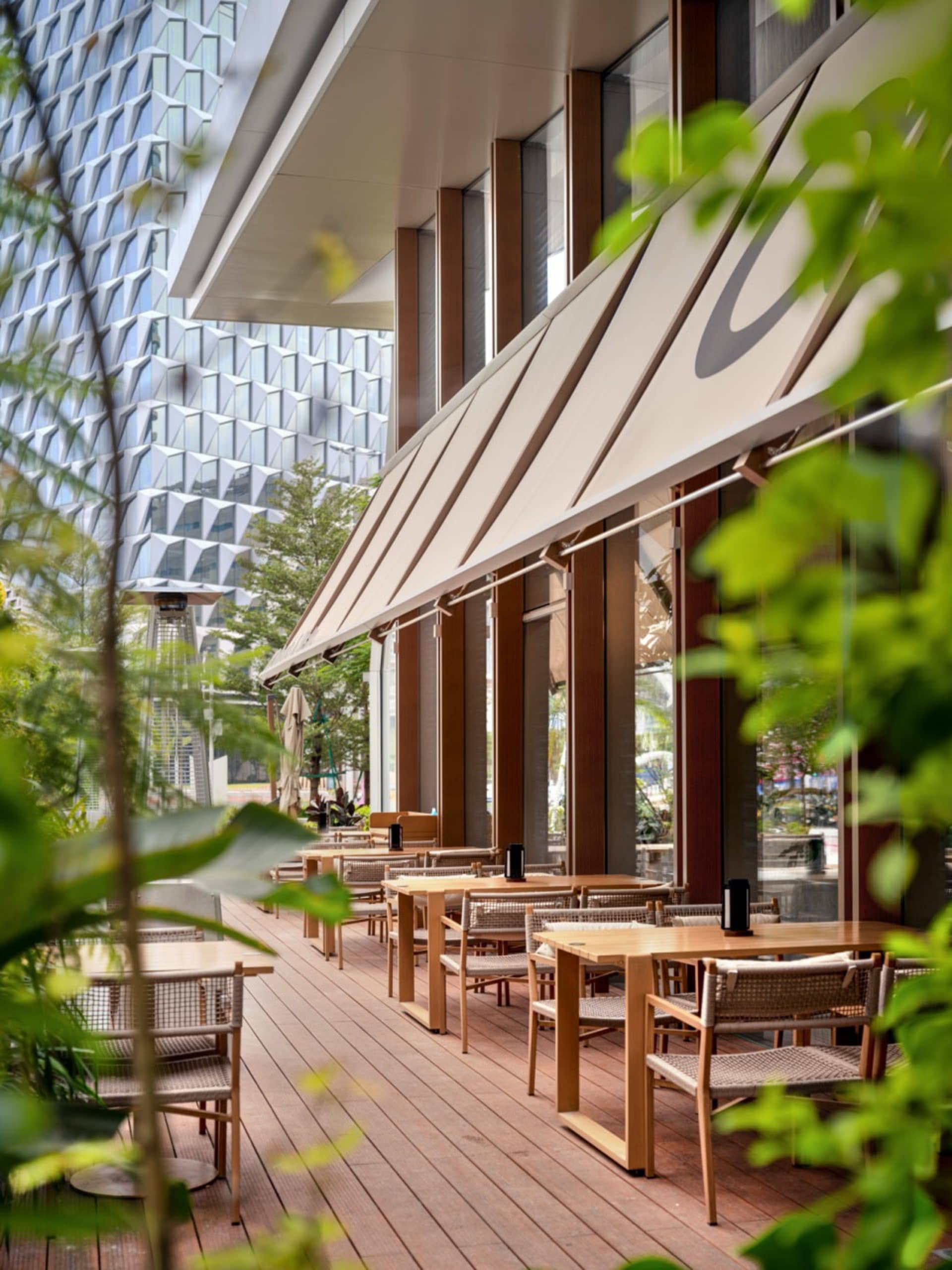 Modern outdoor restaurant patio featuring teak wood tables and chairs with woven seats, beige retractable awning, wooden deck, and lush green plants in foreground with city buildings in background.