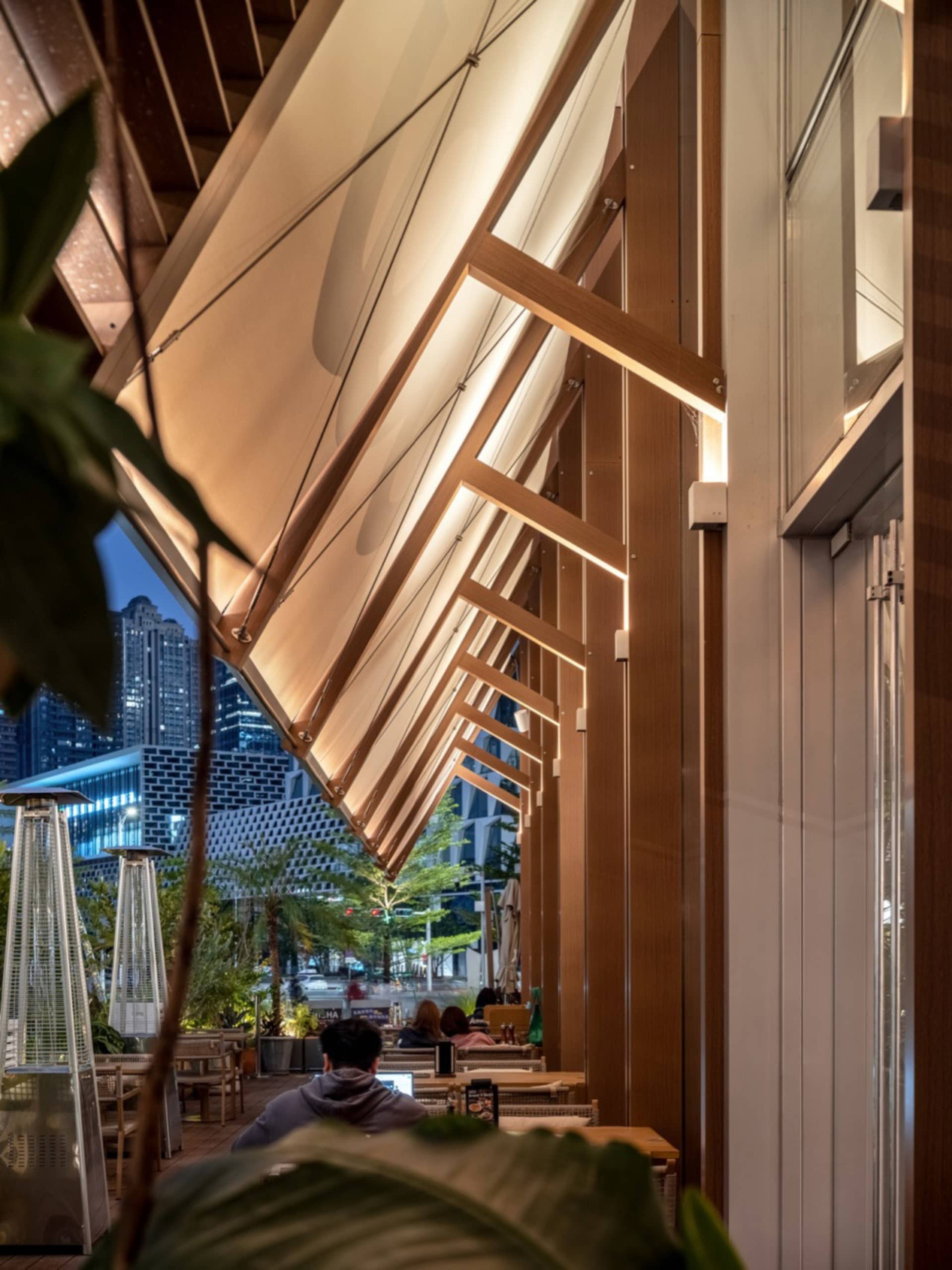 Night view of an upscale outdoor restaurant terrace with a dramatic wooden slat pergola, integrated linear LED lighting, patio heaters, and city skyline in the background.