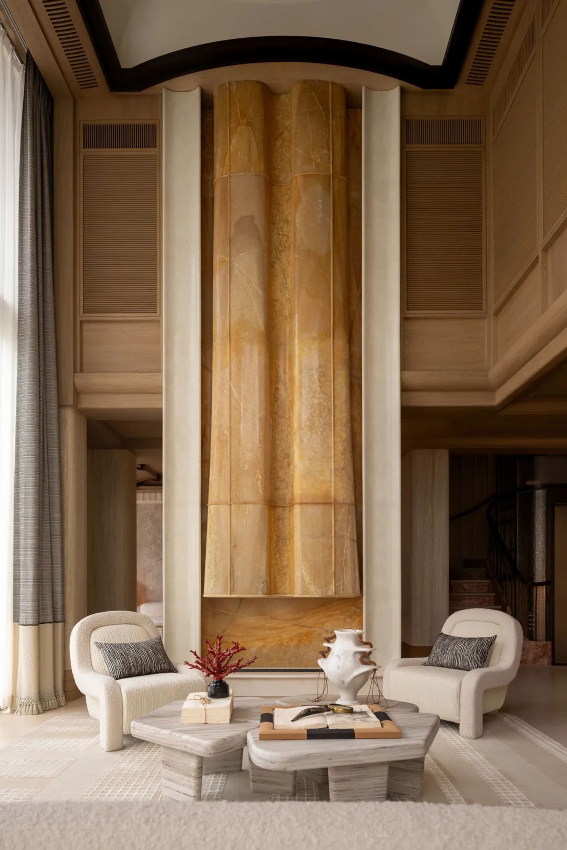 Modern luxury living room featuring a tall translucent golden onyx monolith flanked by two curved bouclé armchairs and travertine coffee tables on a striped rug.
