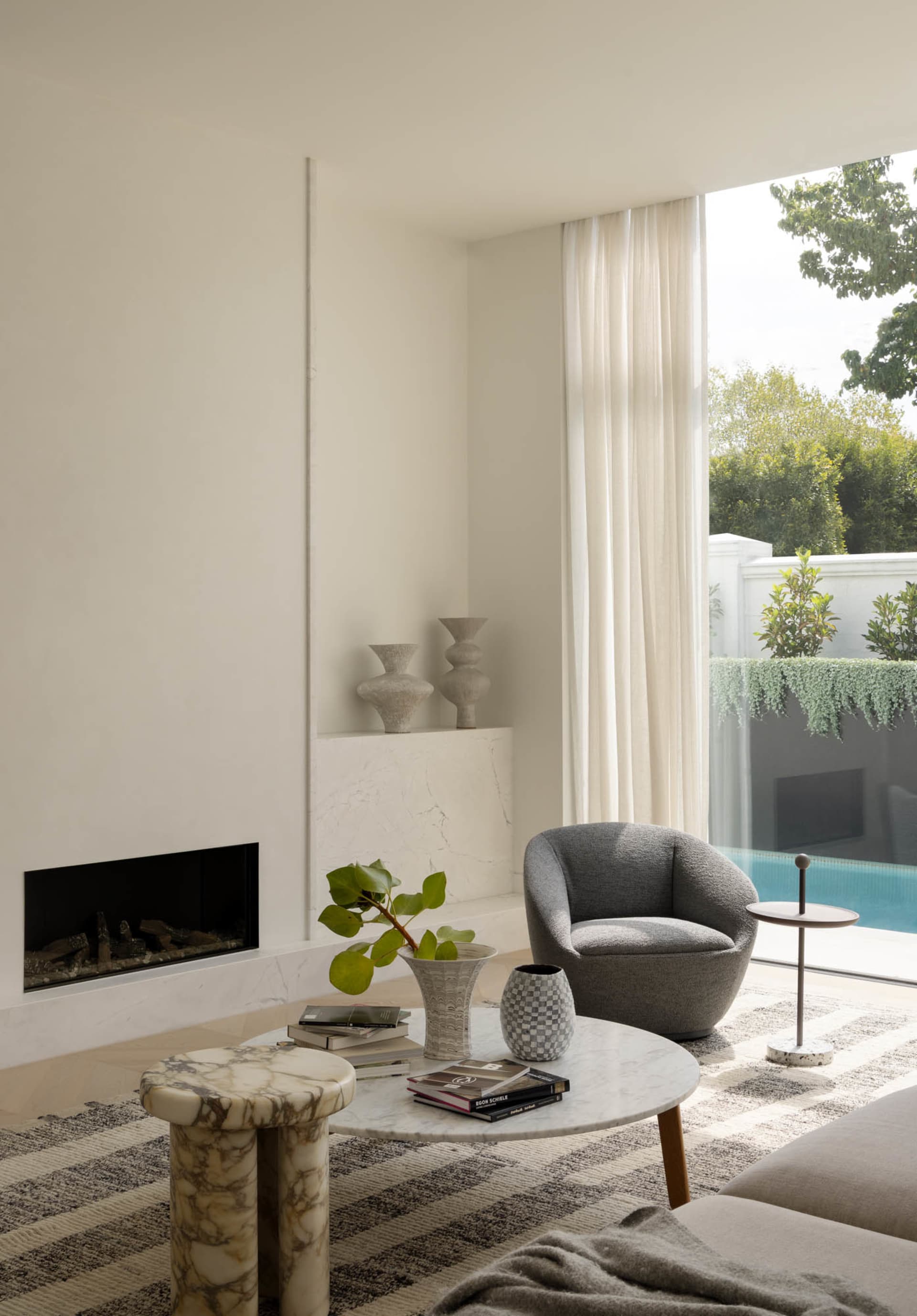 Minimalist living room featuring a marble fireplace, curved grey bouclé armchair, round marble coffee table, and large glass doors opening to a swimming pool and garden.