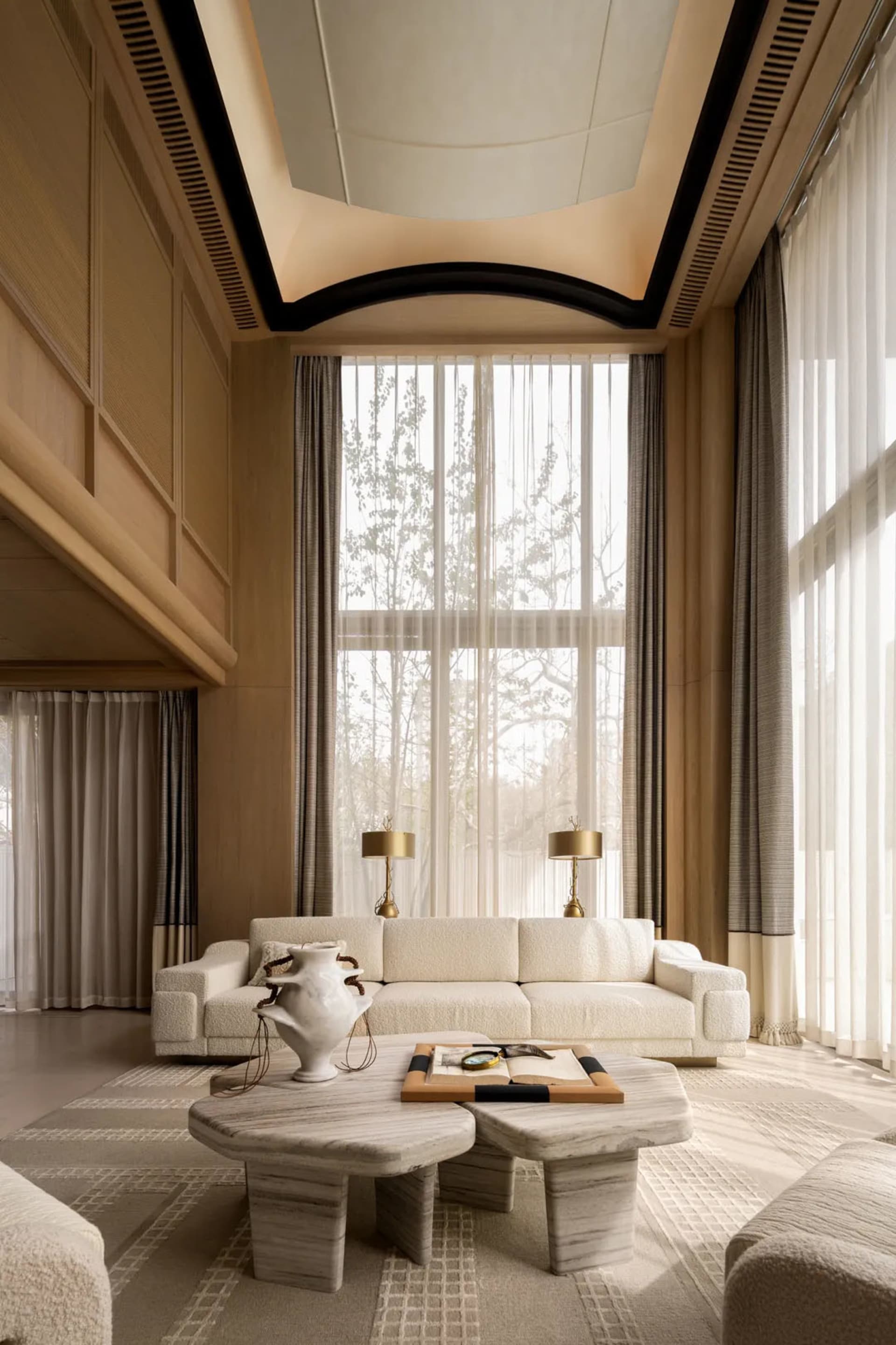 Modern minimalist living room with double-height curved ceiling, off-white bouclé sofa, travertine coffee tables, and floor-to-ceiling sheer curtains allowing natural daylight.