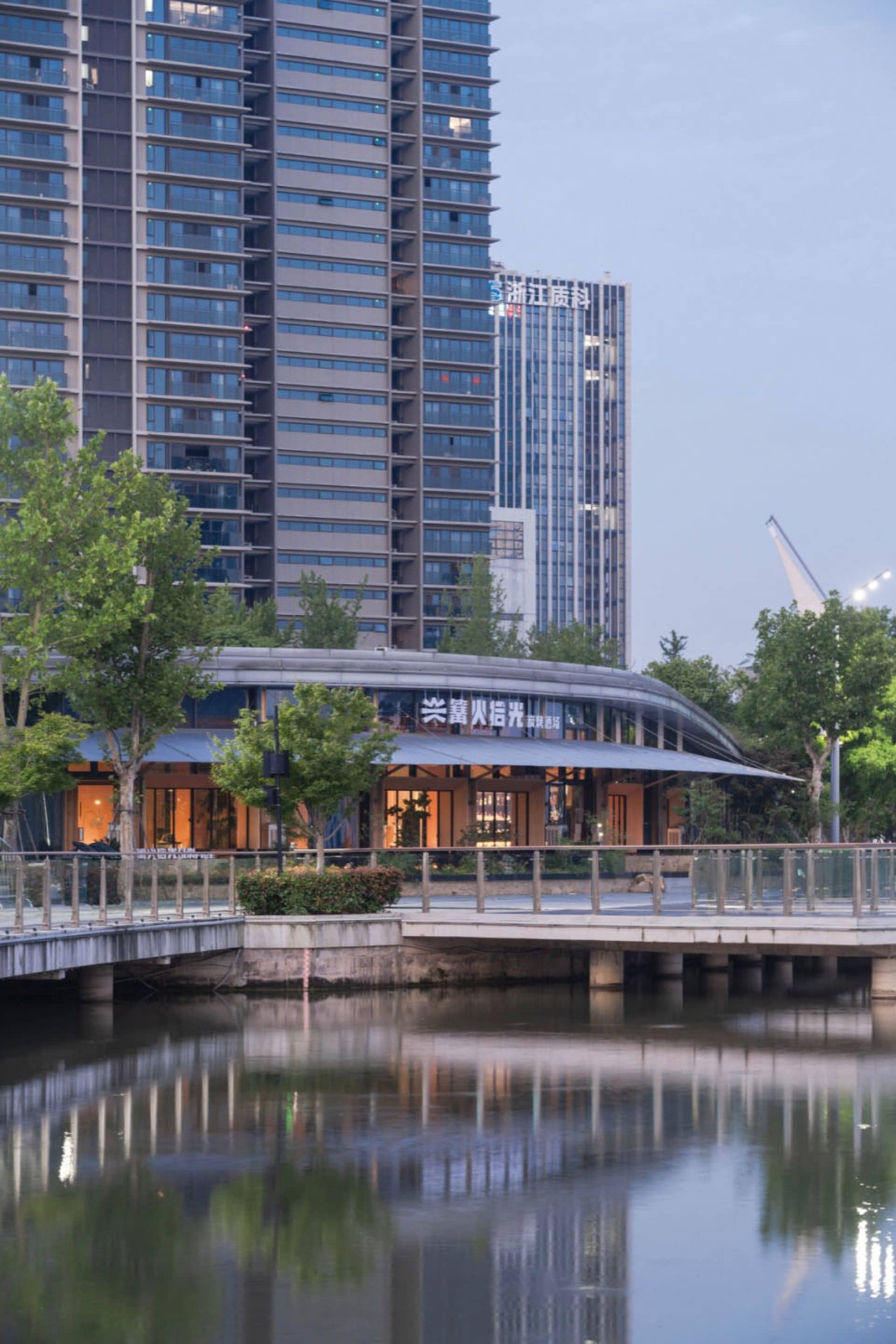 Modern curved waterfront pavilion with illuminated glass facade reflected in a pond at dusk, with high-rise buildings in the background.