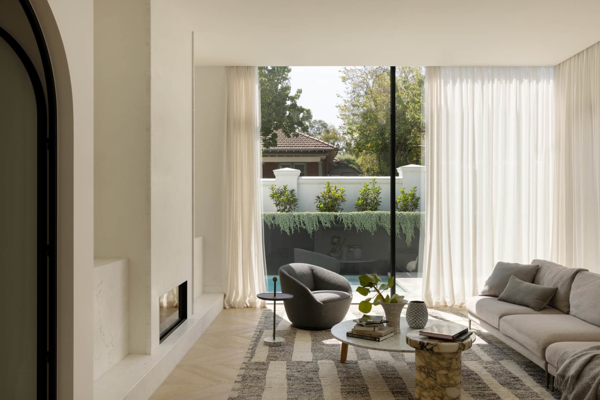 Modern minimalist living room featuring a built-in marble fireplace, light grey sofa, curved grey armchair, marble coffee tables, and large glass doors opening to a swimming pool and garden.
