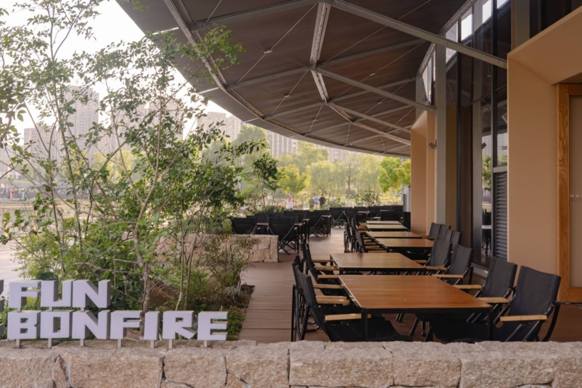 Modern outdoor restaurant patio with wooden tables, black metal chairs, stone retaining wall, and lush greenery under a curved fabric canopy during daytime.