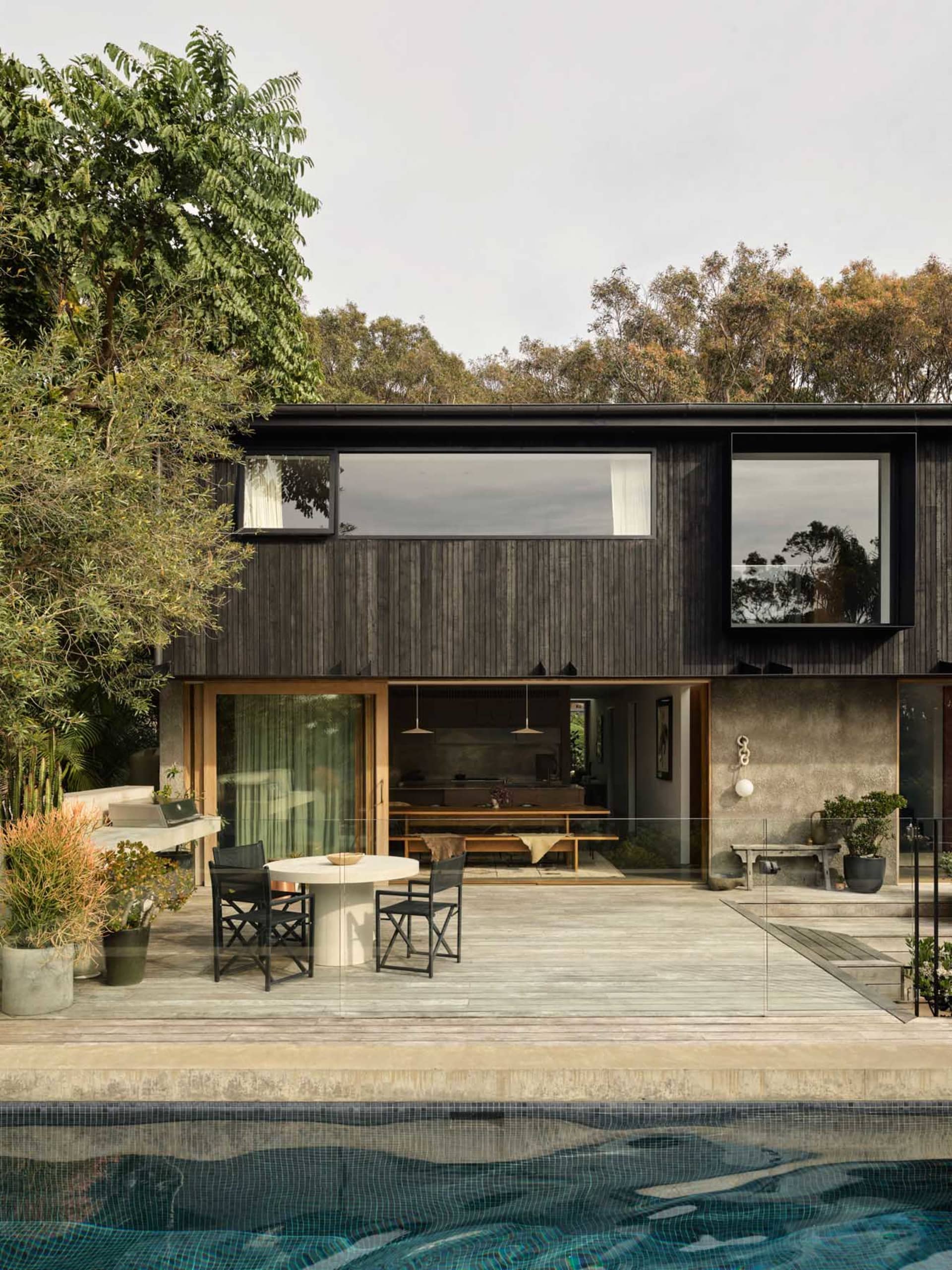 Modern two-story villa with dark charred wood facade, large glass windows, outdoor dining area with round table and black chairs, and a swimming pool in the foreground.