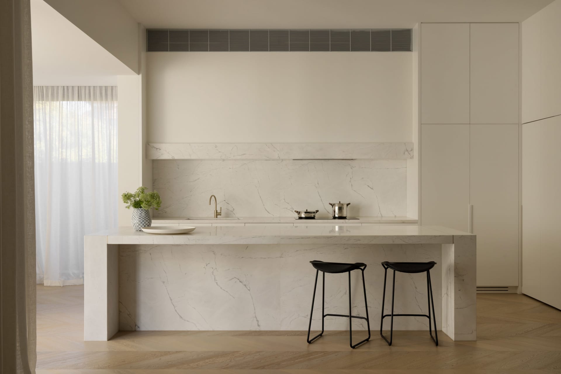 Modern minimalist kitchen featuring a large white marble island with two black wireframe stools, matching marble backsplash and counter, brass faucet, and soft natural light from a window with sheer curtains.