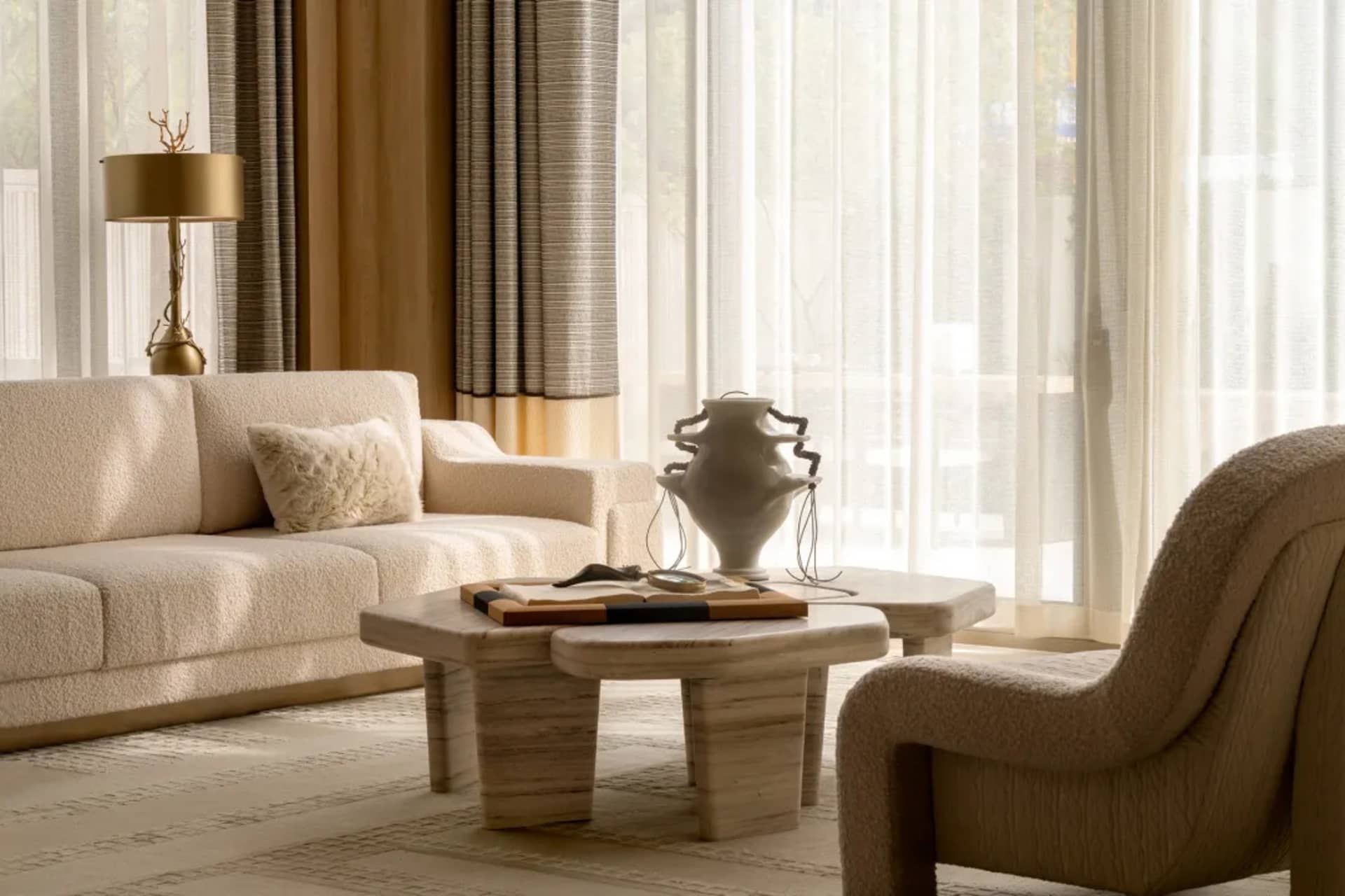 Bright minimalist living room featuring a cream boucle sofa, travertine coffee table, sculptural white vase, and sheer curtains with warm neutral tones.