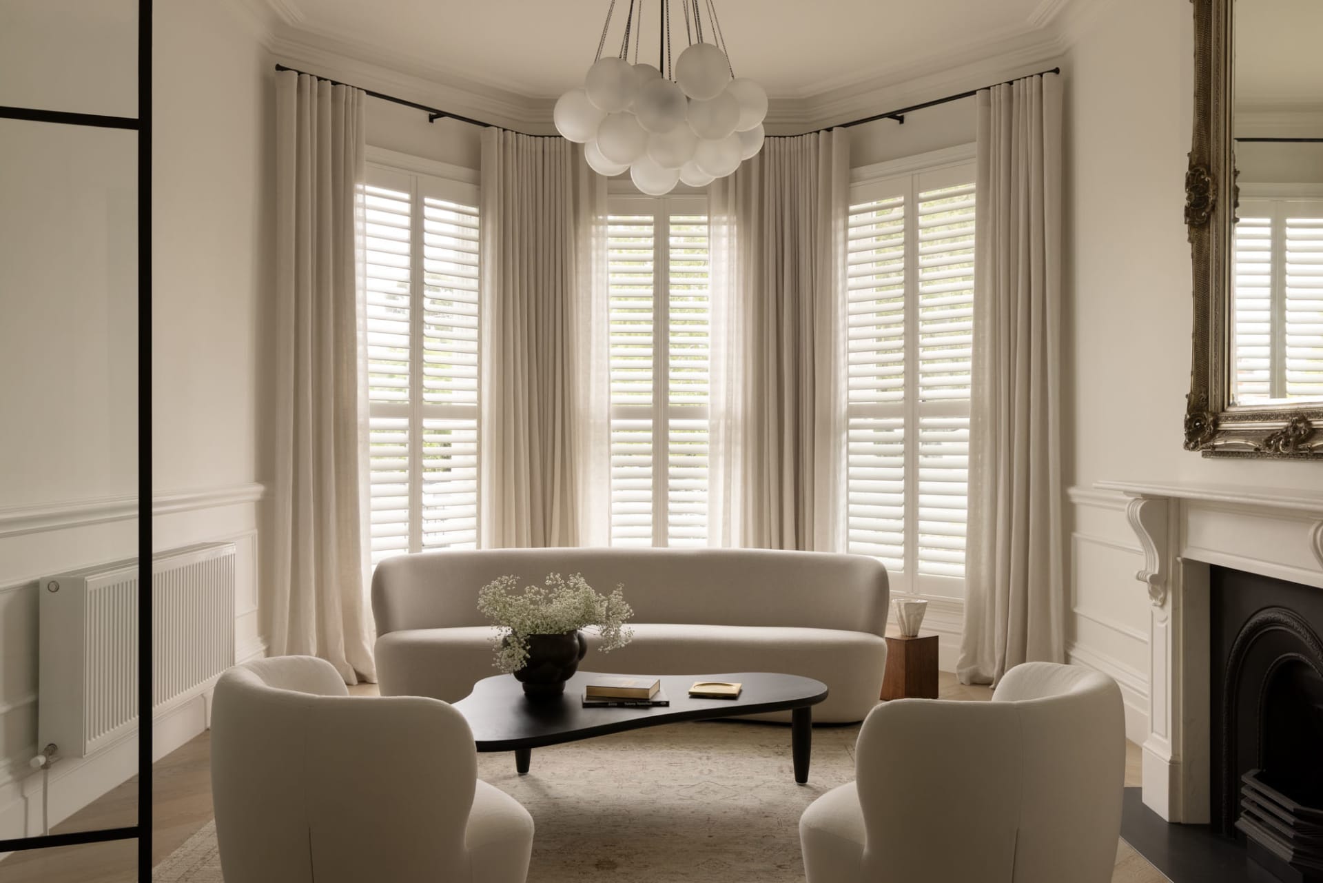 Bright and airy living room featuring a curved boucle sofa, two matching armchairs, a black organic coffee table, and a cloud-like chandelier in front of a large bay window with white shutters and curtains.