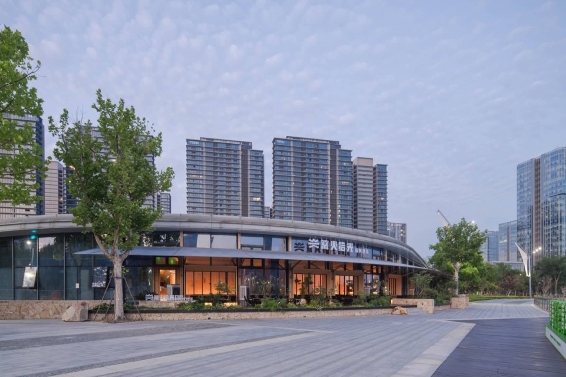 Modern curved glass and concrete community center with illuminated interior, set in a spacious urban plaza with trees and high-rise residential buildings in the background at dusk.