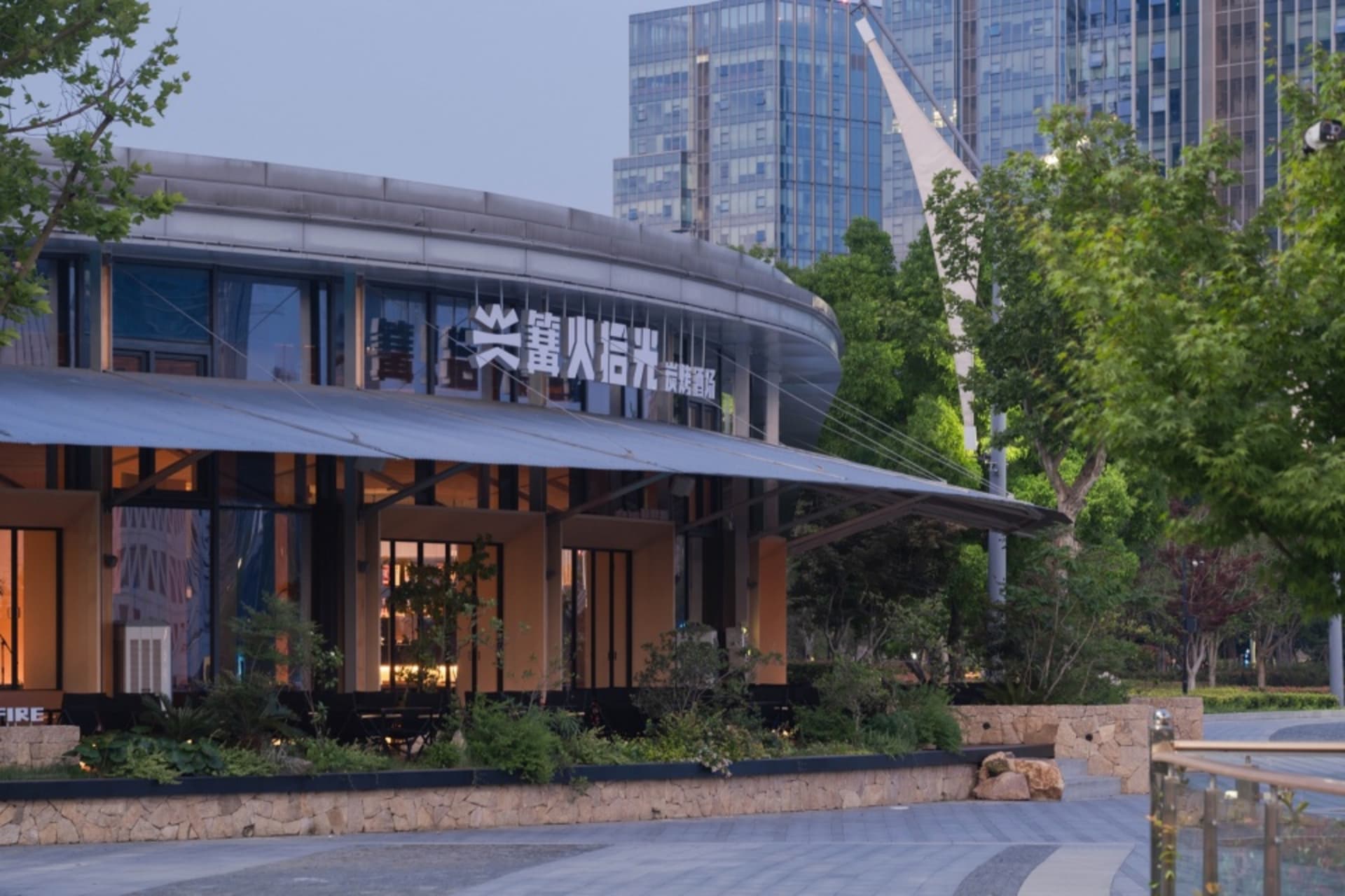 Modern curved restaurant exterior with stone base, large glass windows, and illuminated signage at dusk, surrounded by trees and city buildings in the background.