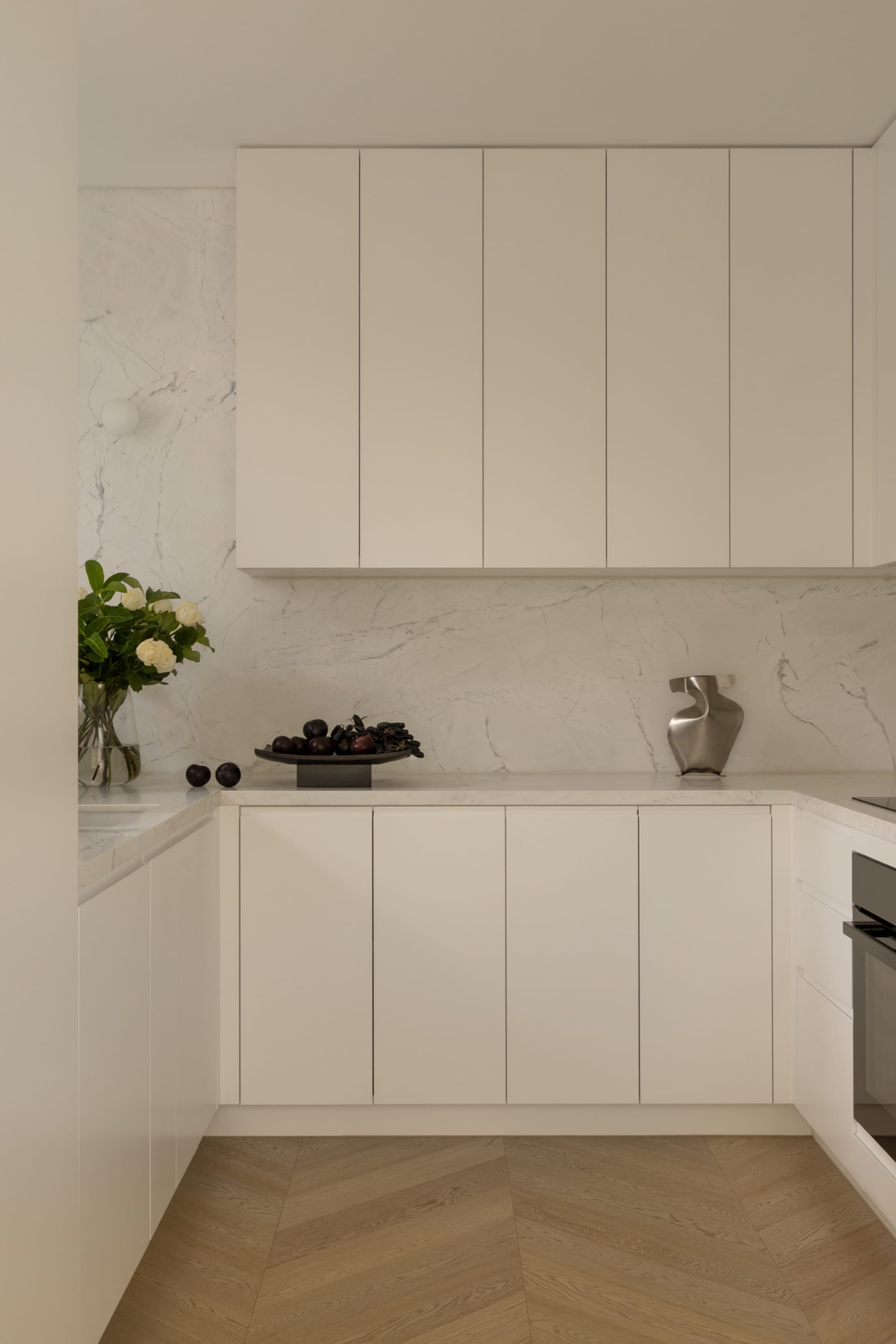 Modern minimalist kitchen featuring white flat-panel cabinetry, Calacatta marble backsplash and countertops, light herringbone wood flooring, and decorative objects including a flower vase and metallic sculpture.