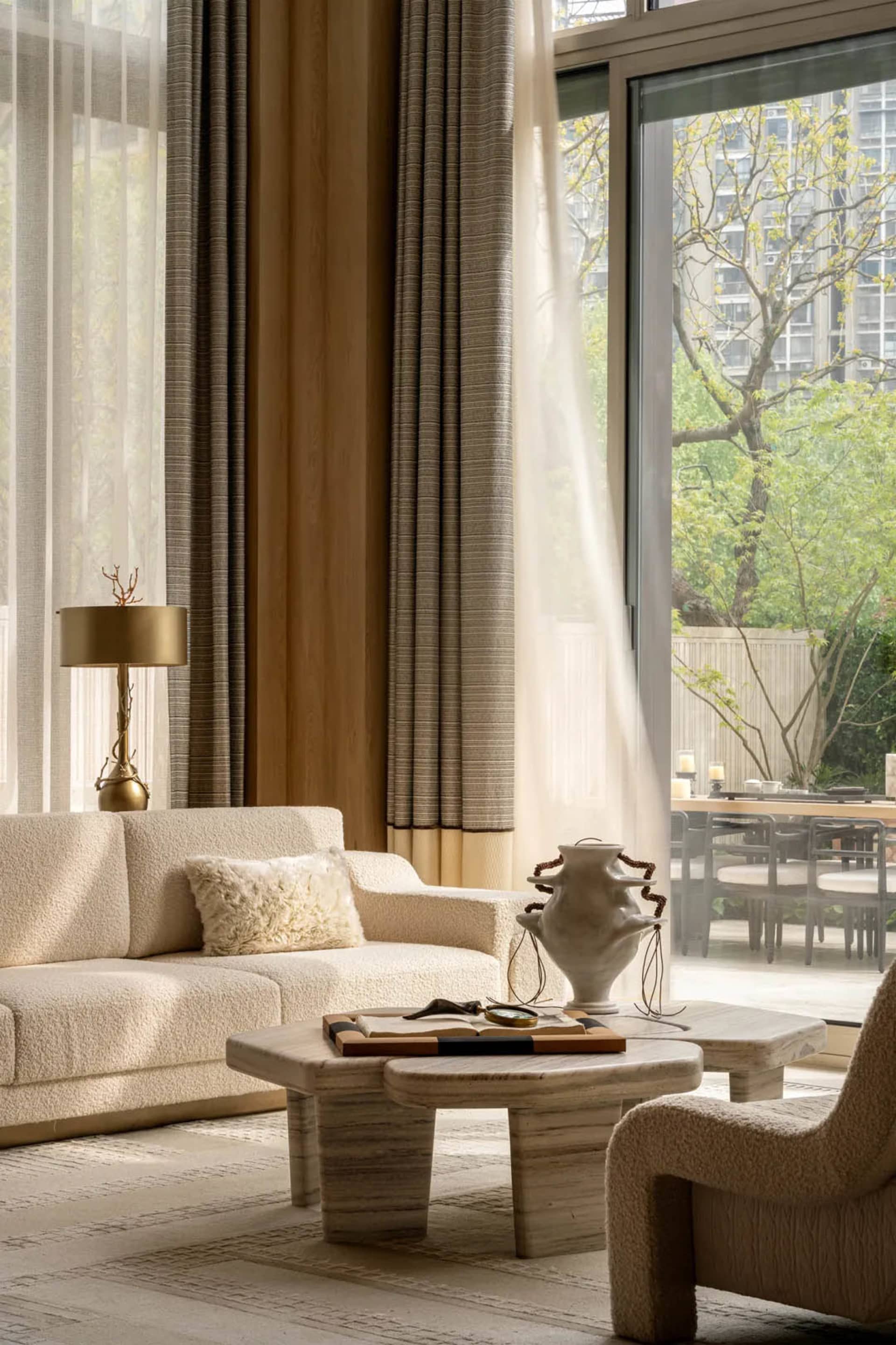 Modern living room featuring a cream boucle sofa, travertine coffee tables, sculptural ceramic vase, and large glass doors opening to a green garden view.