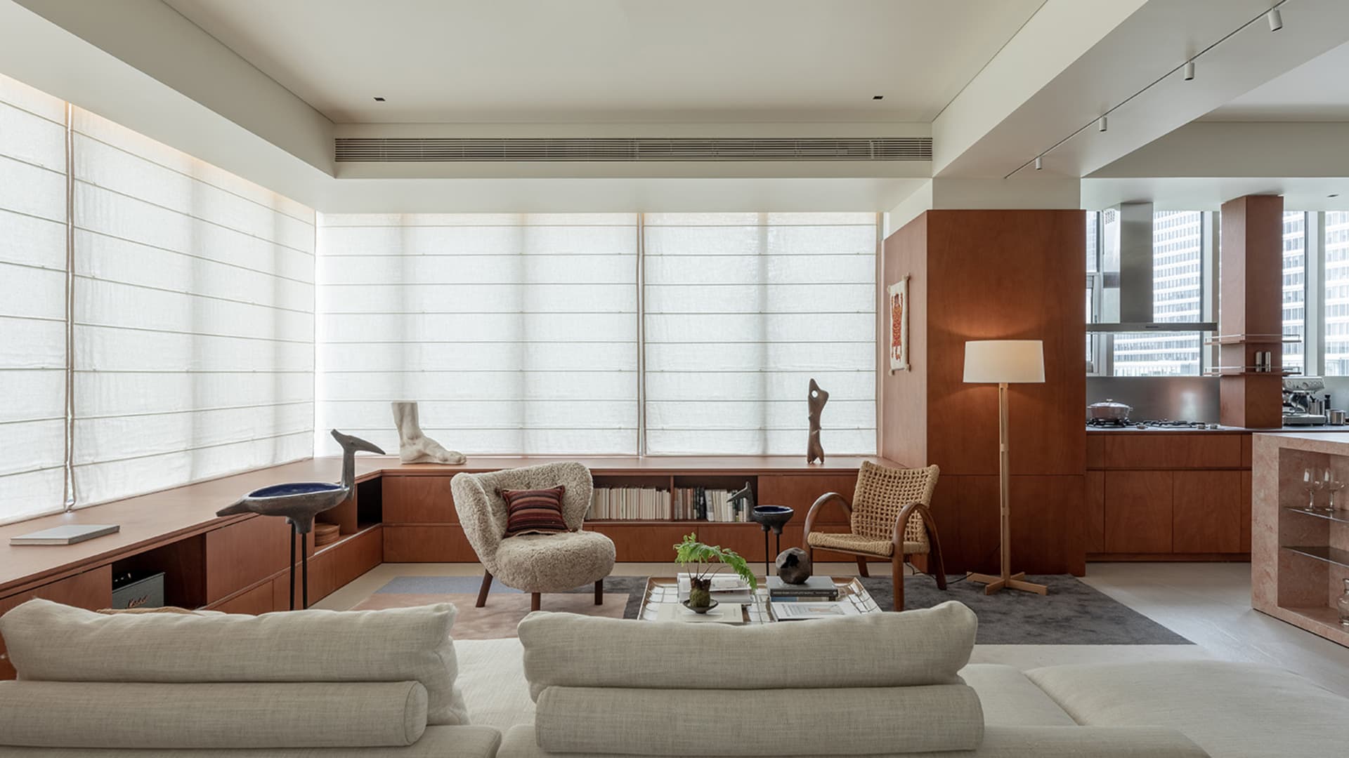 Modern minimalist living room featuring extensive walnut built-in cabinetry, a large boucle armchair, a rattan lounge chair, and a light-colored sectional sofa with large windows covered by sheer roman shades.