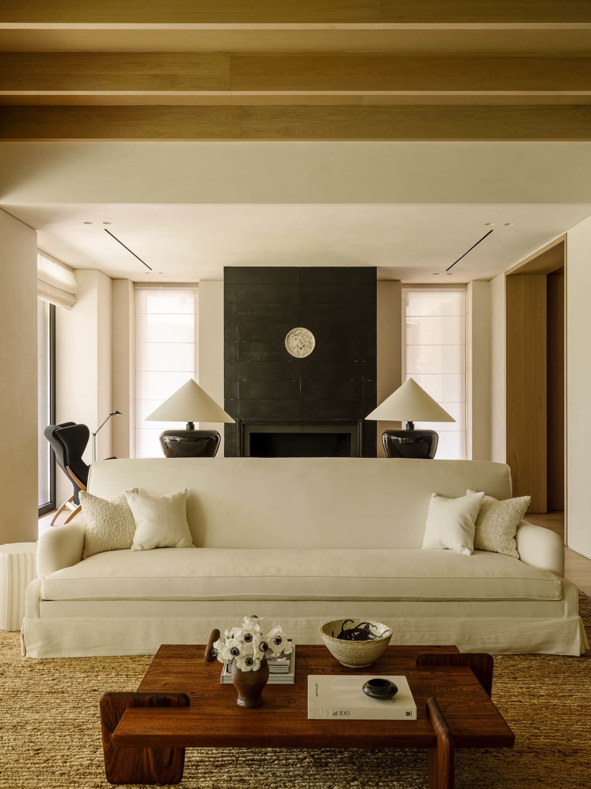 Modern minimalist living room featuring a large off-white sofa, dark walnut coffee table, black stone fireplace, and warm wooden ceiling beams.