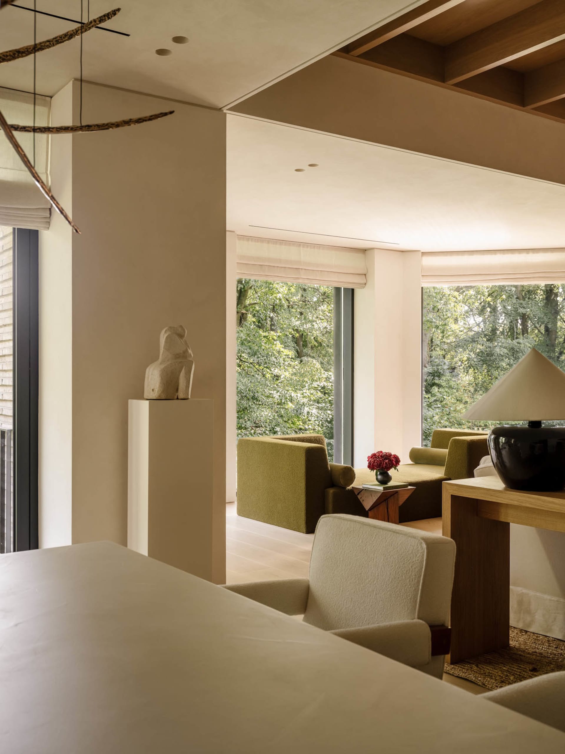 Modern living room interior featuring olive green sofas, light oak furniture, a stone sculpture on a pedestal, large windows overlooking a lush forest, and a black ceramic table lamp.