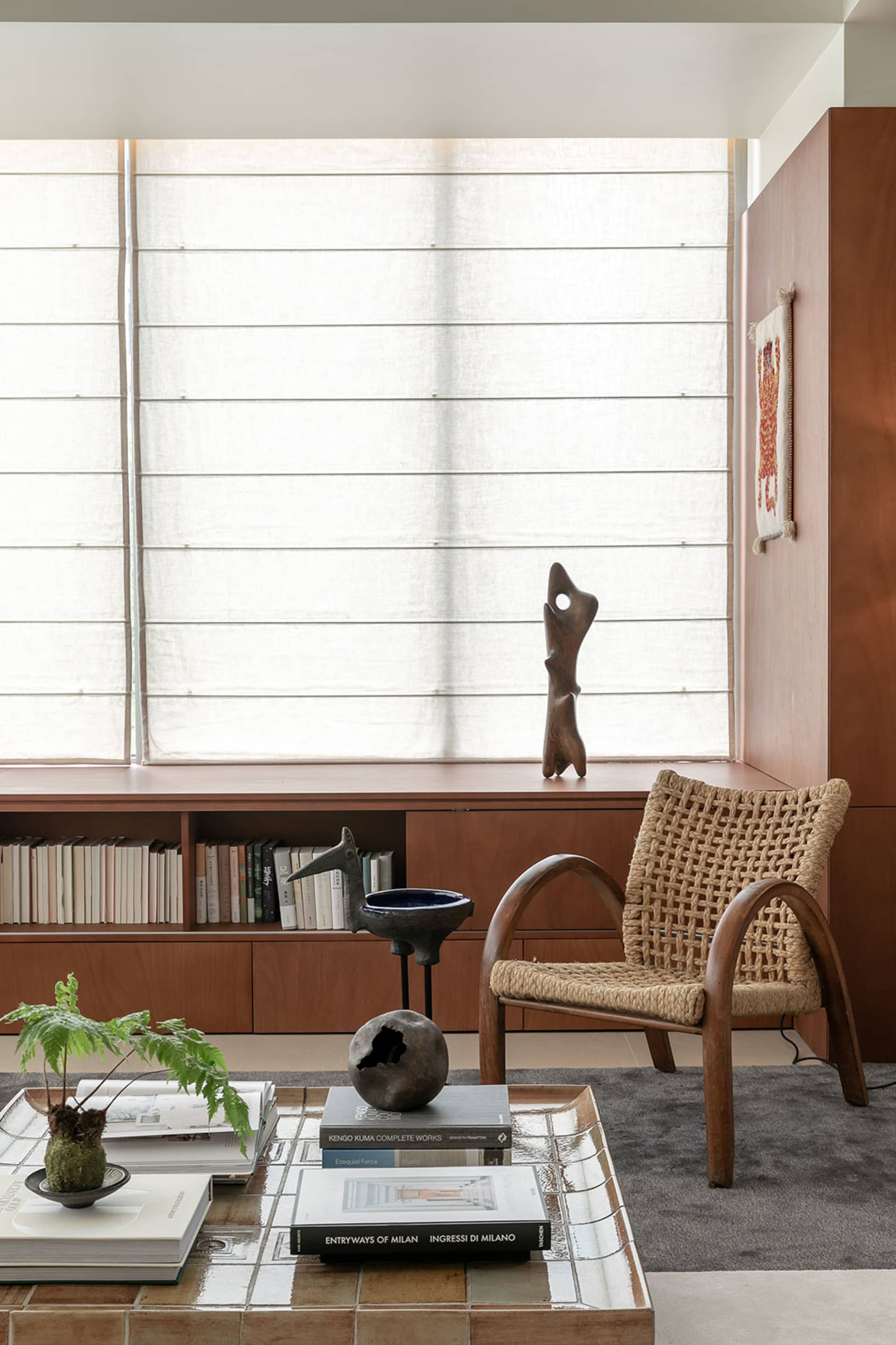 Modern minimalist living room featuring a wooden rope-weave armchair, built-in teak window seat with linen roman shades, ceramic sculptures, and books on a tiled coffee table.