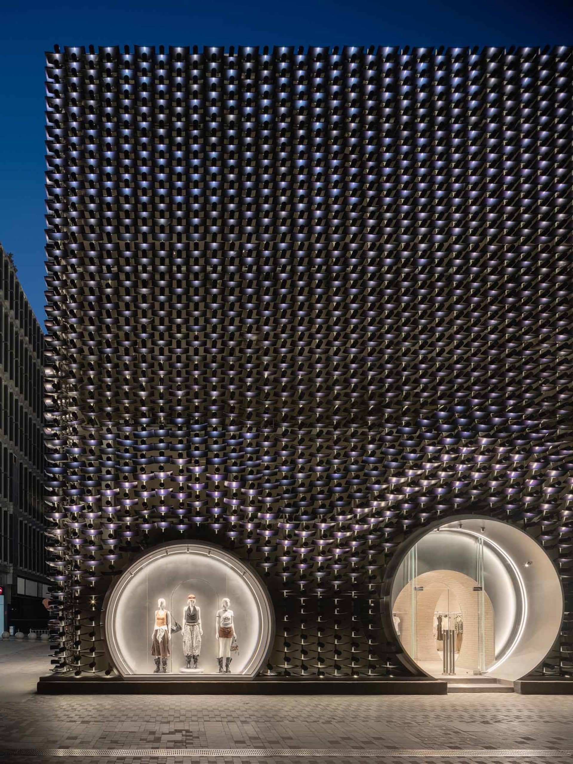 Exterior night view of a luxury retail building featuring a dramatic black perforated metal facade with two large illuminated circular entrances displaying mannequins.