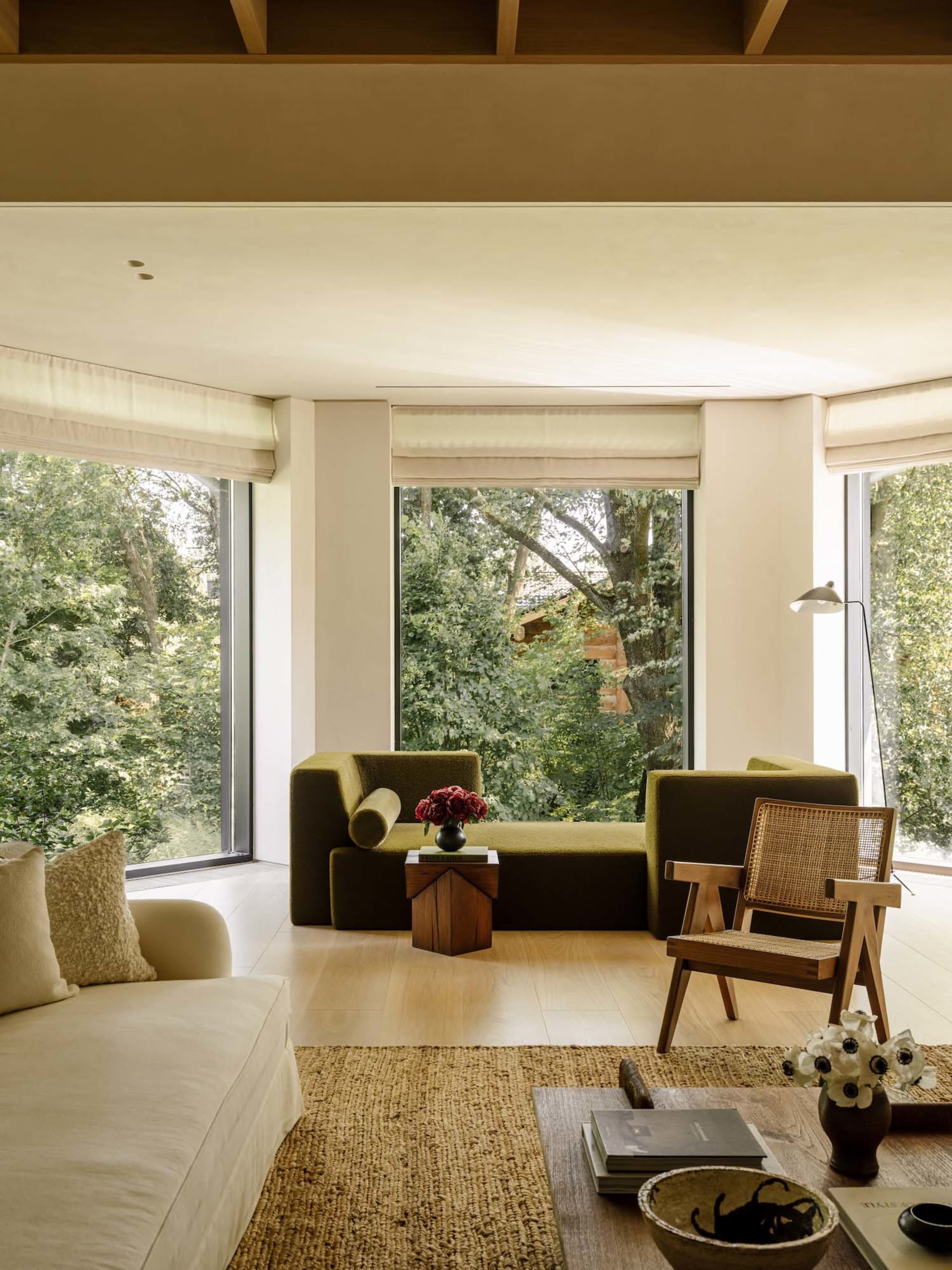 Modern living room featuring an olive green boucle daybed, wooden armchair with cane back, cream sofa, and large windows overlooking lush green trees.