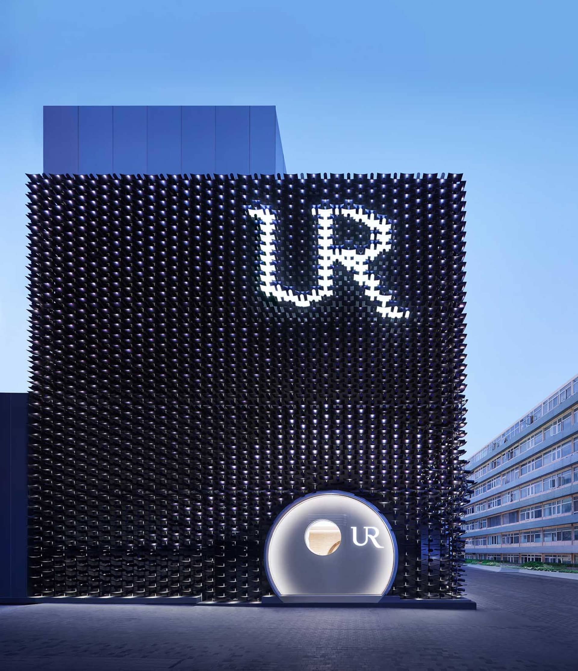 Night view of a modern building with a black perforated metal facade featuring a large illuminated UR logo and a circular entrance structure.