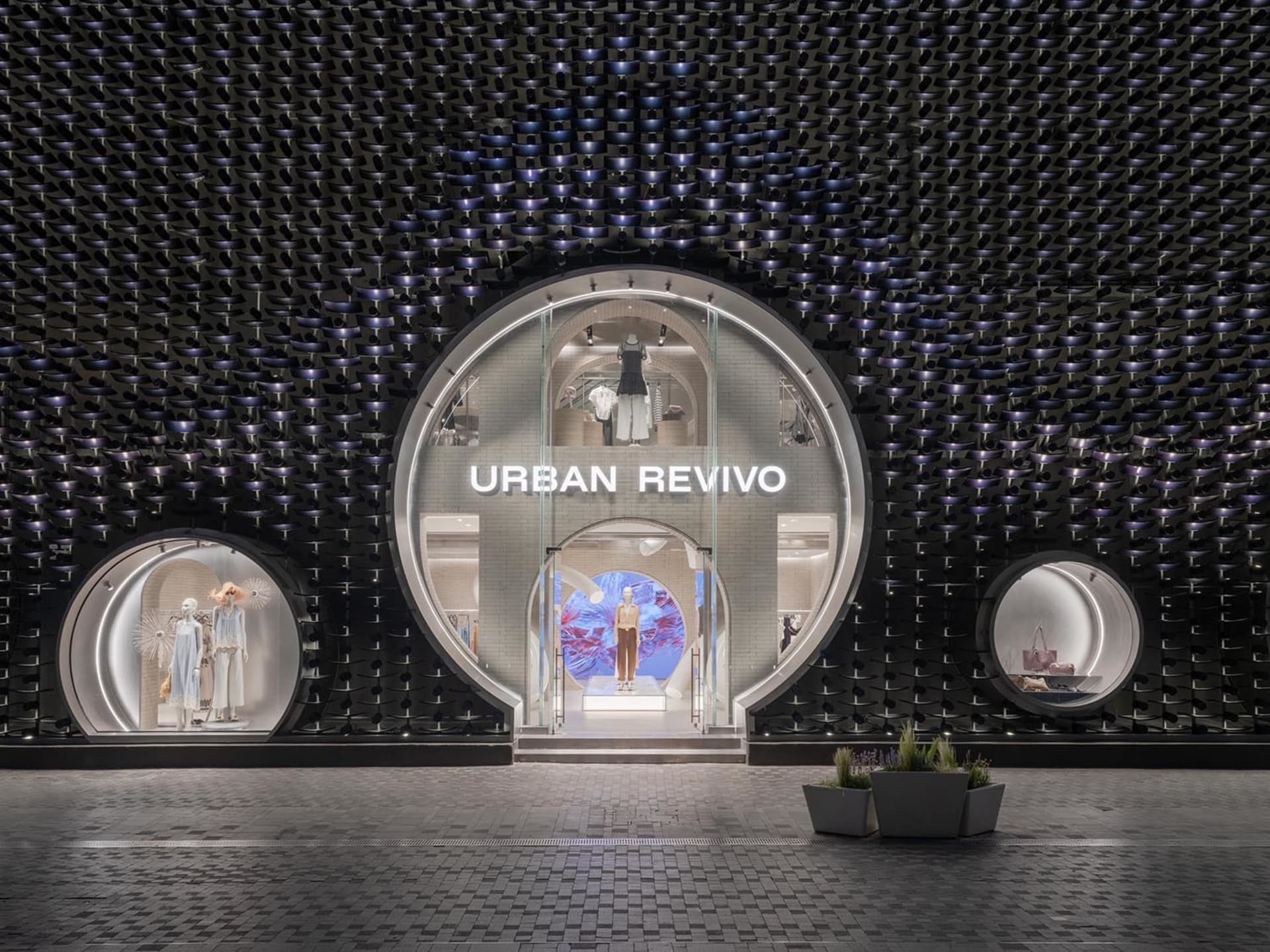 Night view of Urban Revivo fashion store featuring a large circular glass entrance set in a dramatic black textured sculptural wall with smaller circular display windows on either side.