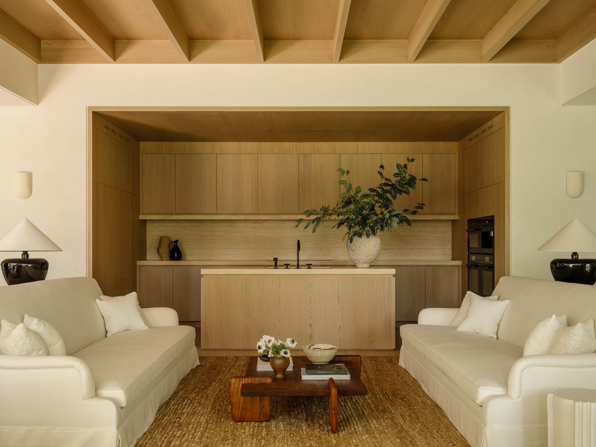 Modern minimalist living room with two white sofas facing a wooden coffee table on a jute rug, opening into a clean white oak kitchen with exposed ceiling beams.