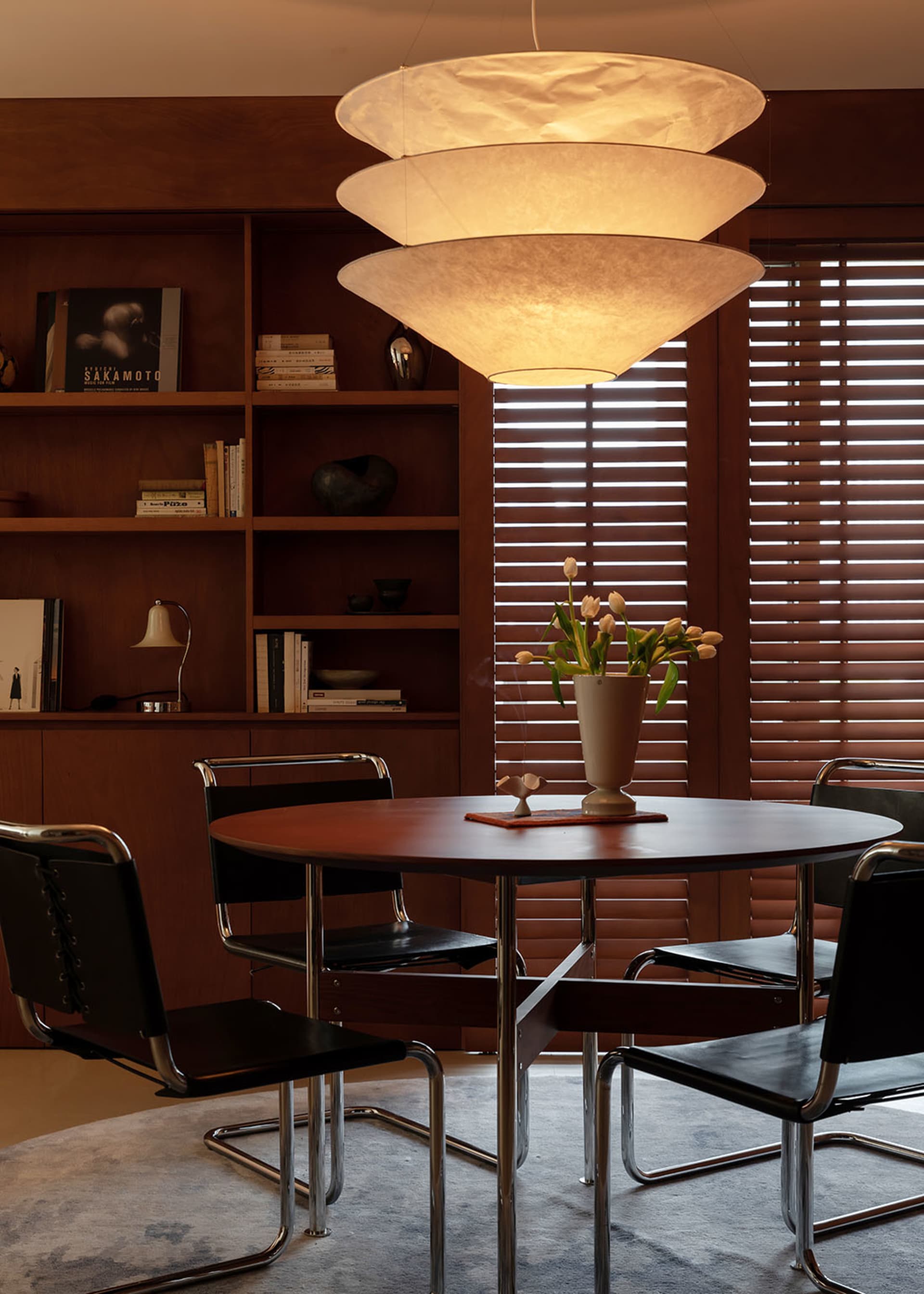 Mid-century modern dining room with round walnut table, black leather cantilever chairs, large tiered fabric pendant light, and built-in walnut shelving.