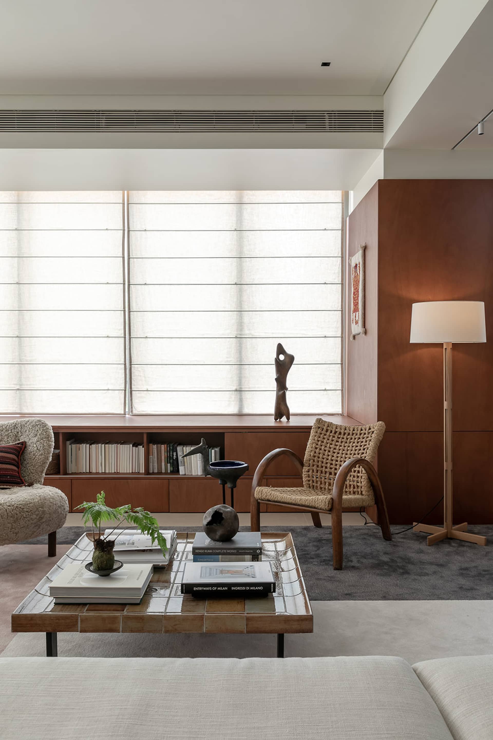 Modern living room featuring teak wood cabinetry, a woven rattan armchair, tiled coffee table, bouclé sofa, and large window with sheer blinds.