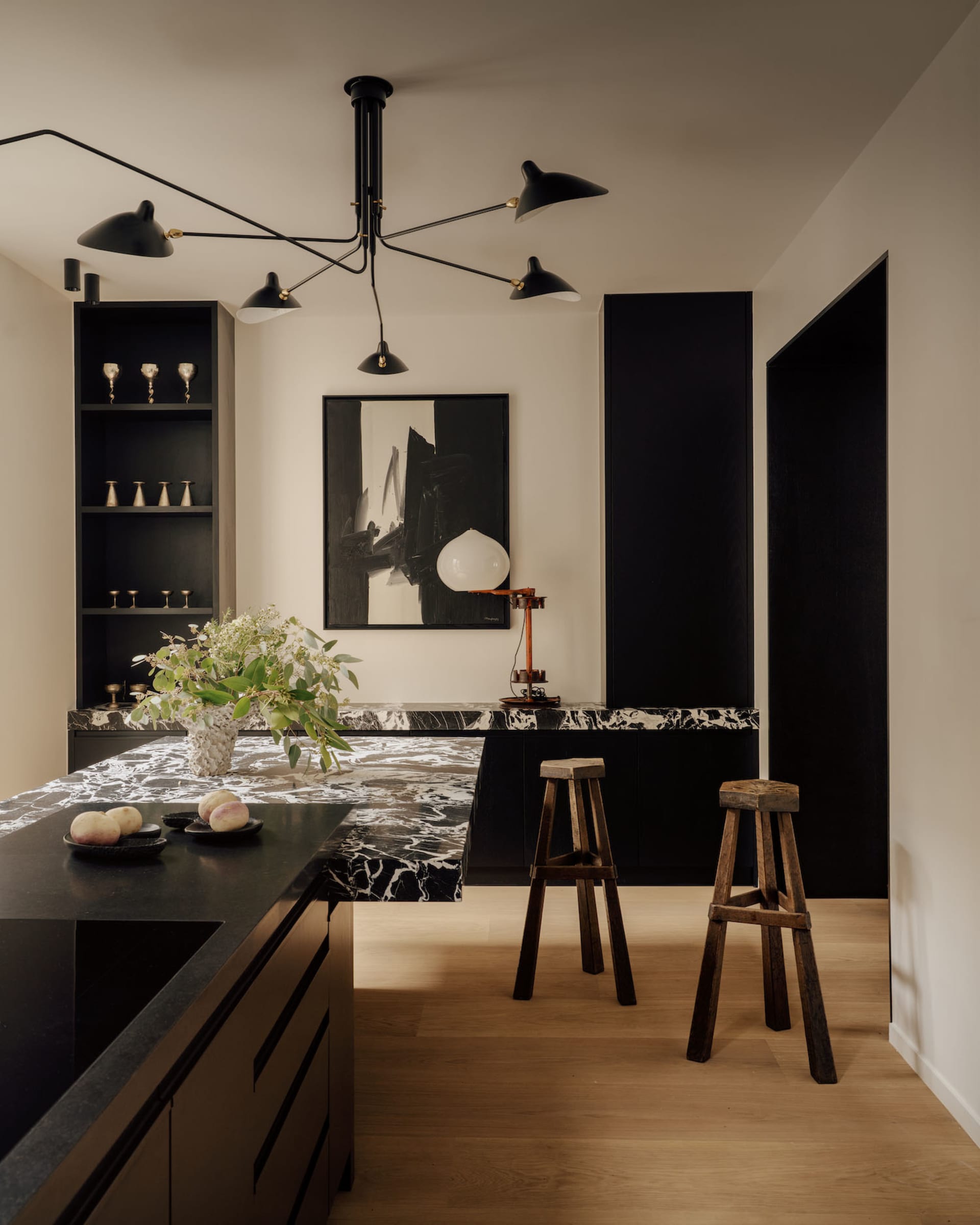Modern kitchen with black marble island, dark cabinetry, two rustic wooden bar stools, large black chandelier, and abstract black and white artwork on the wall.