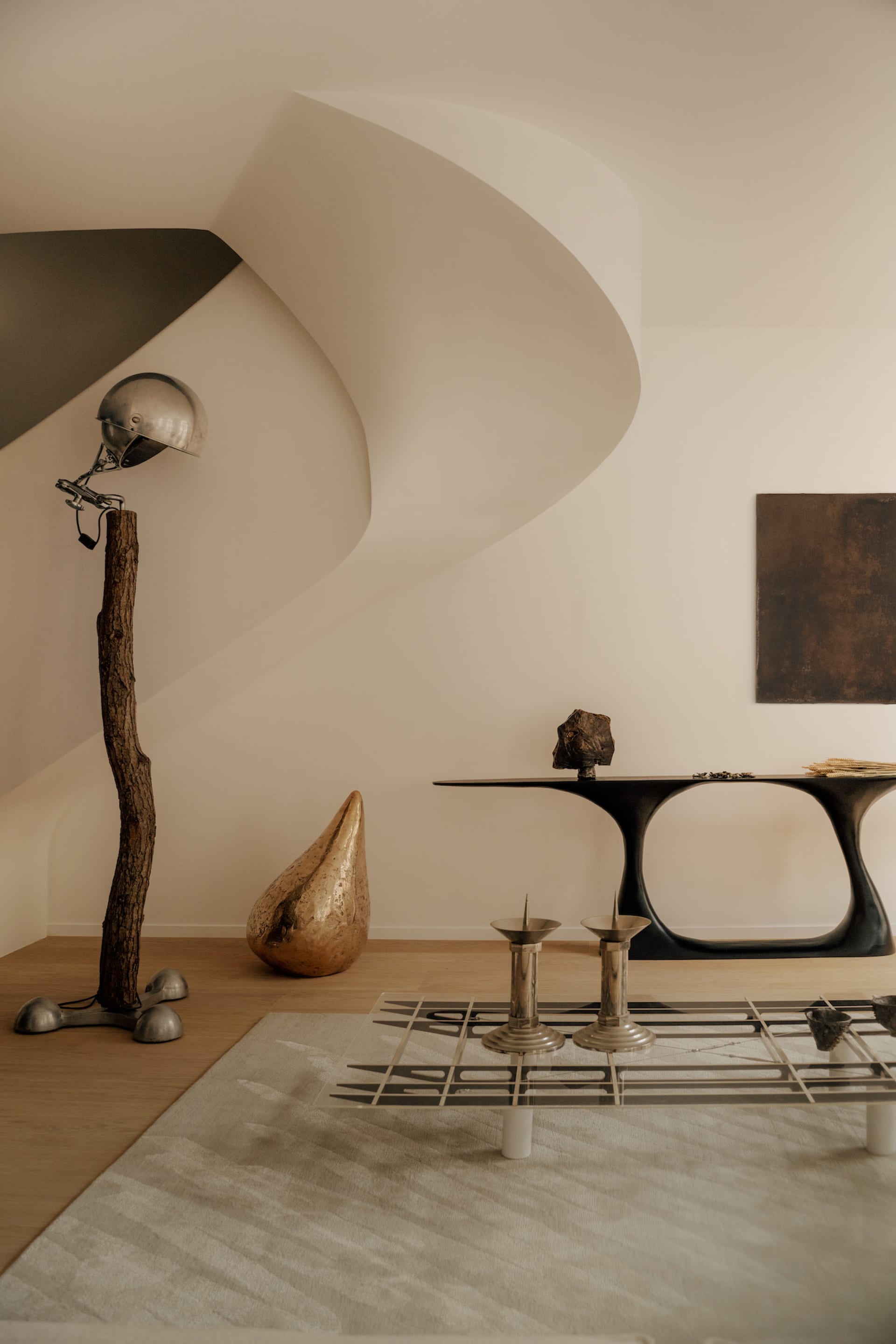 Modern minimalist living room featuring a sculptural black console table, a floor lamp made from a real tree trunk with a vintage helmet shade, a textured gold teardrop sculpture, and a glass coffee table with metallic candle holders on a neutral rug.