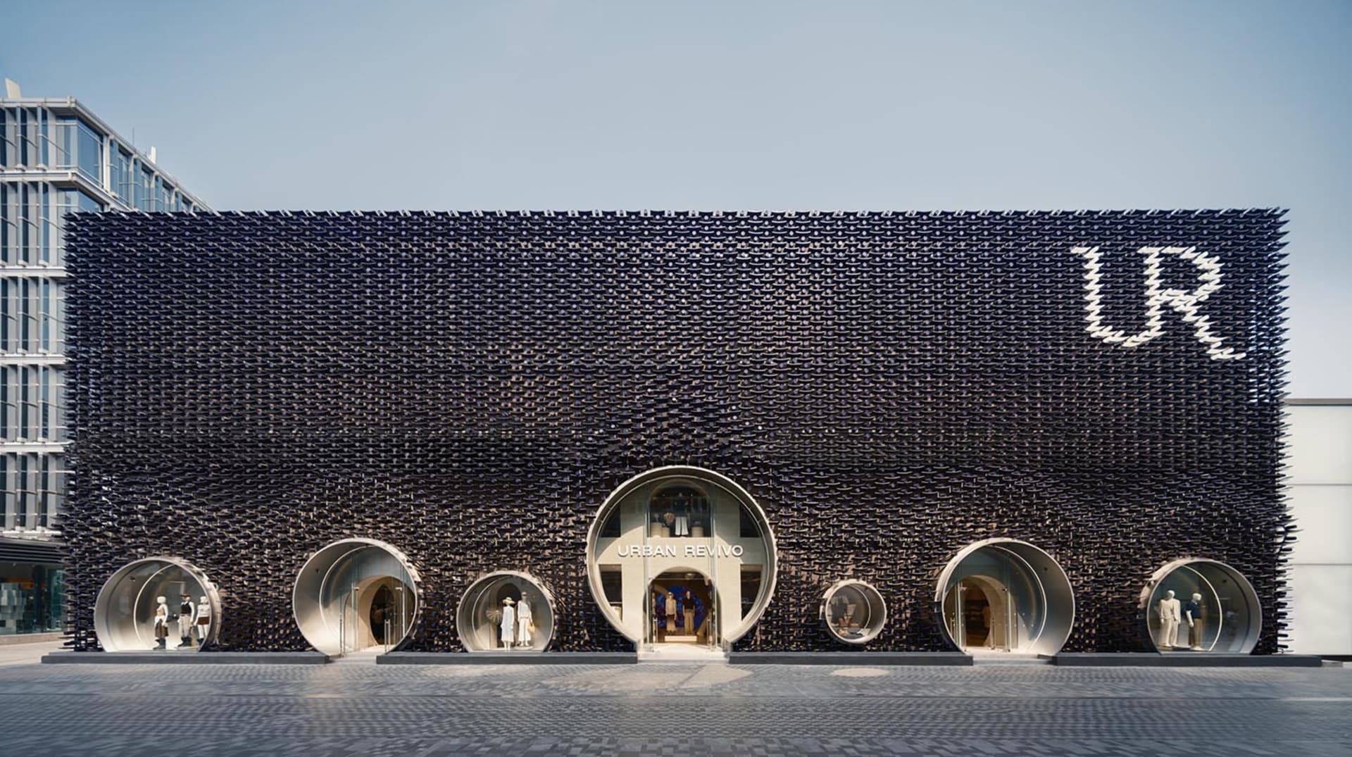 Front view of Urban Revivo flagship store in Taipei featuring a massive black textured façade made of recycled plastic tiles and multiple circular tube-shaped entrances with mannequins displayed inside.