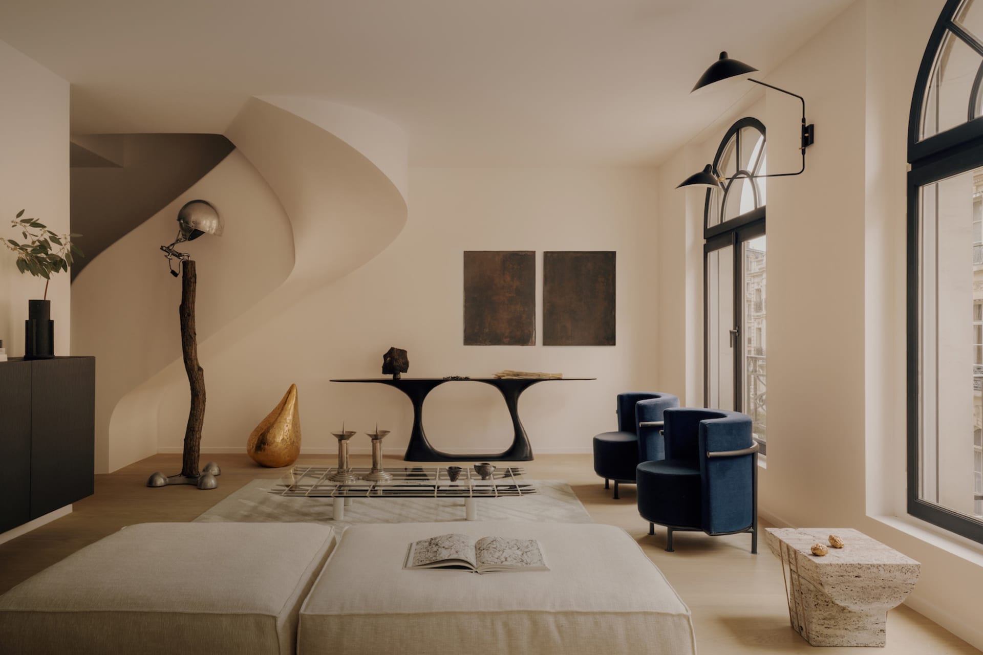 Modern organic living room featuring two large beige floor cushions, blue velvet armchairs, a black sculptural console table, a dramatic curved staircase, and a mix of natural materials including travertine and polished metal.