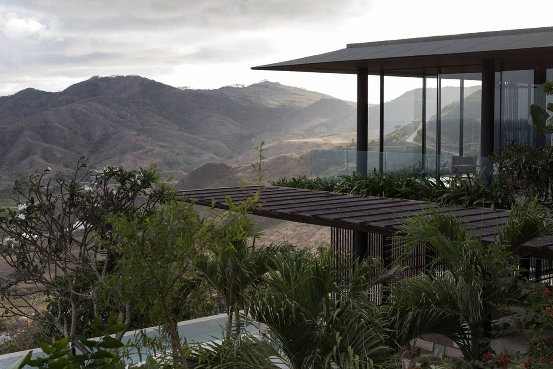 Modern mountain villa with large glass windows and a dark wooden terrace overlooking a vast tropical mountain valley under cloudy skies.