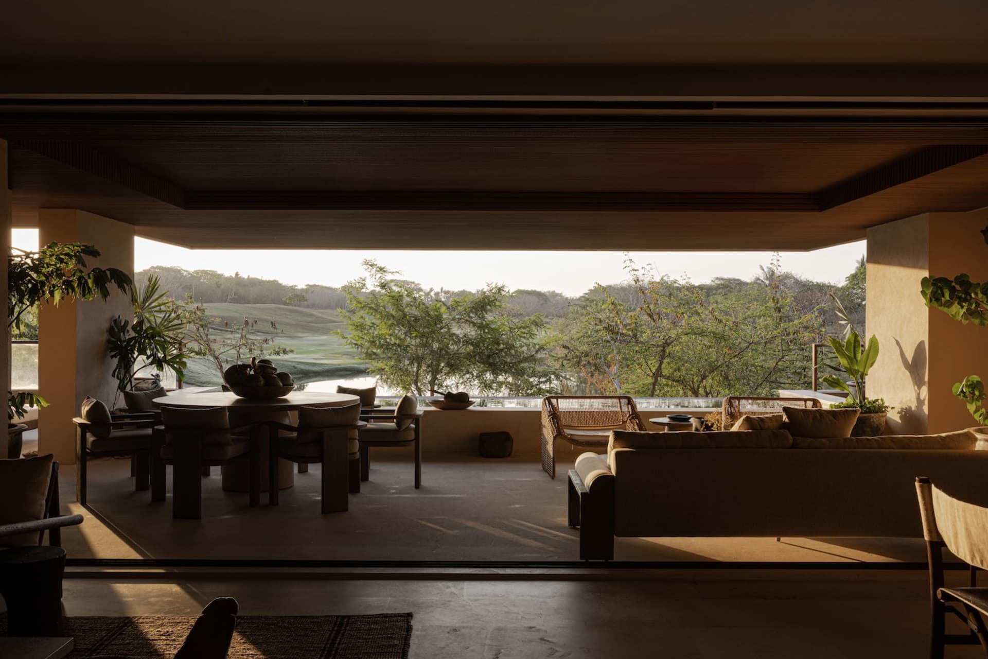 Modern open-plan living and dining area with large sofa, round dining table, and expansive view of a golf course and tropical landscape.