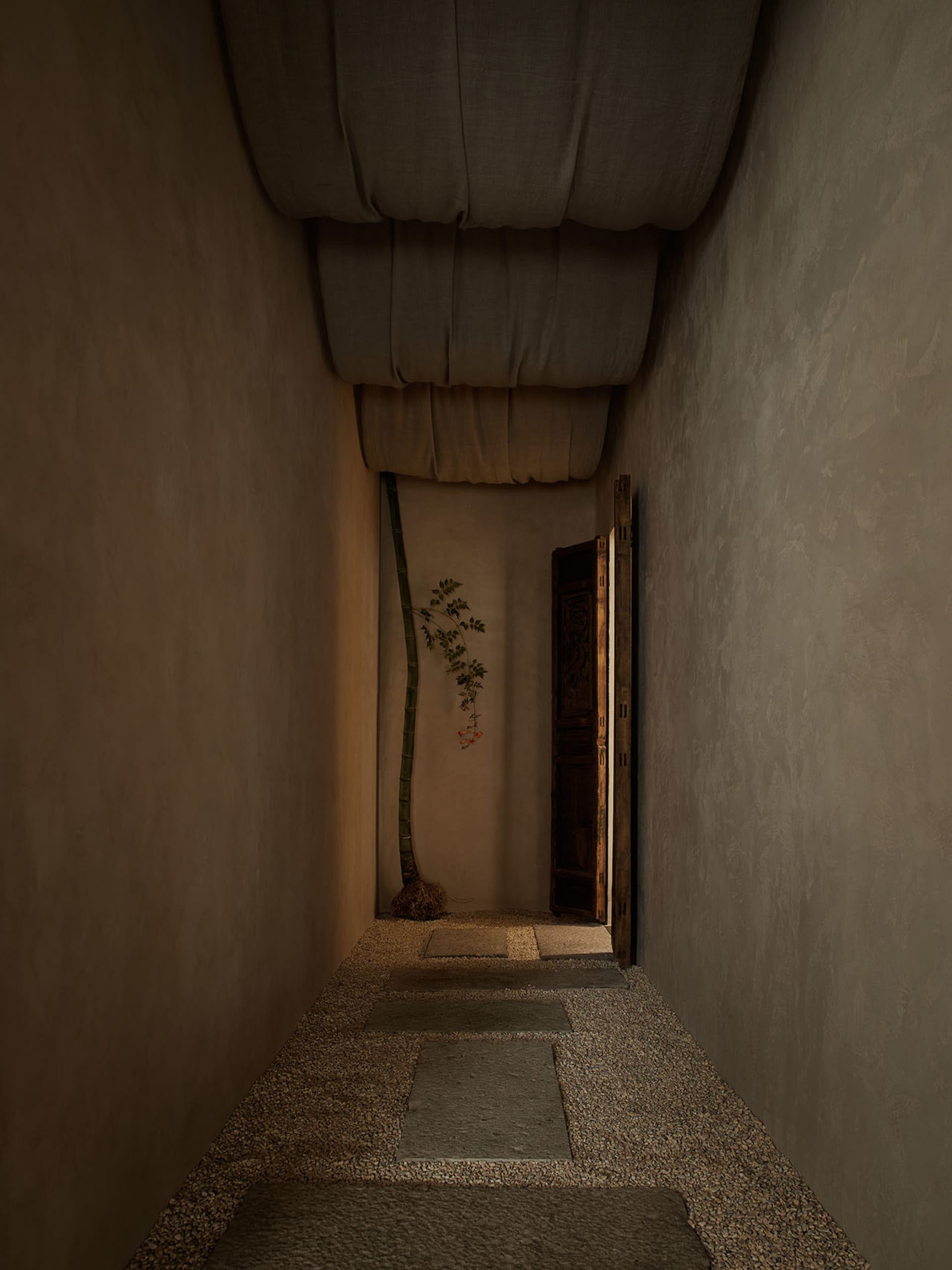 Narrow indoor hallway with textured plaster walls, gravel floor, stepping stone path, a tall bamboo plant, and a carved wooden door at the end.