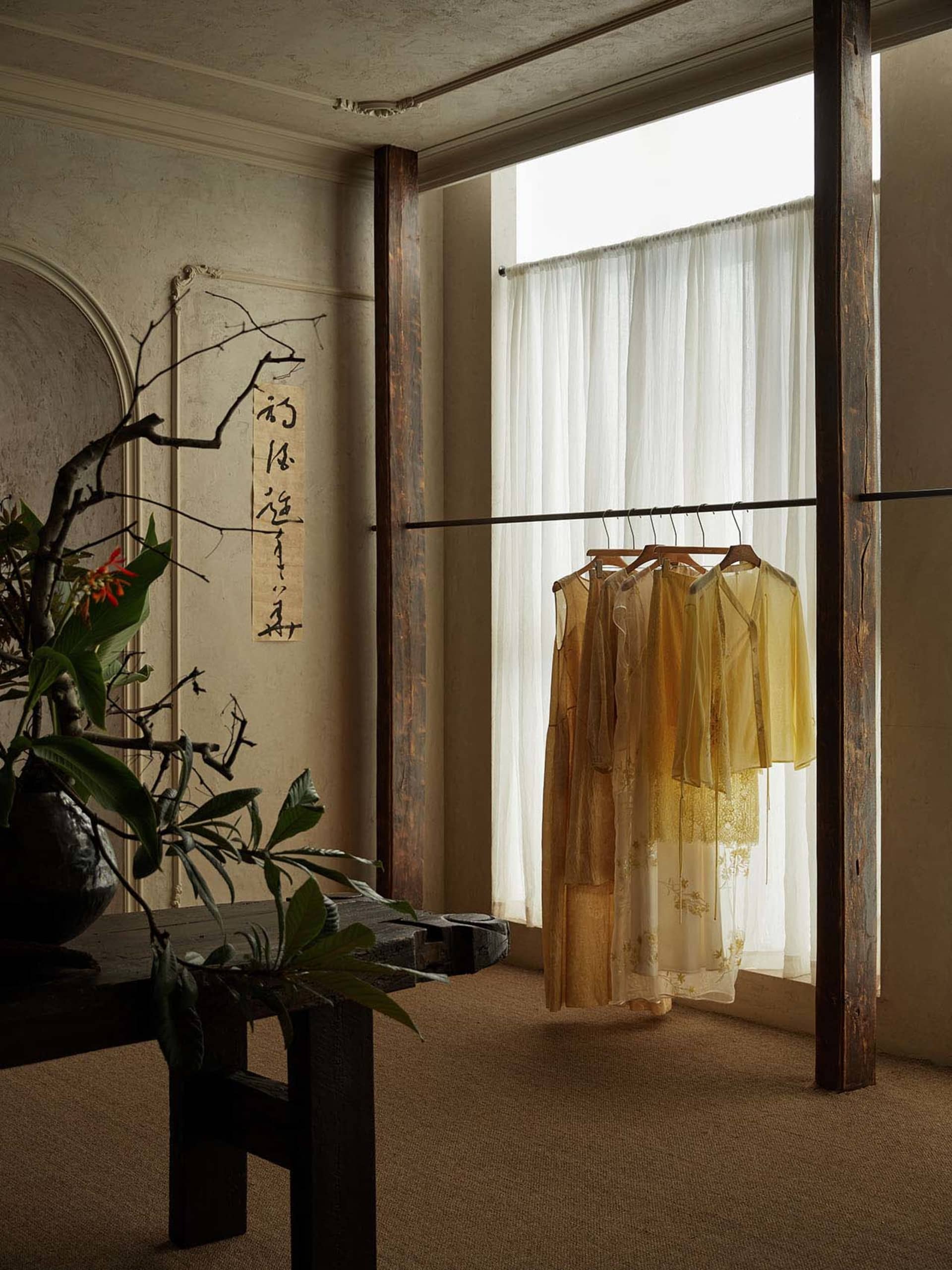 Wabi-sabi style interior corner featuring a dark reclaimed wood clothing rack with hanging yellow and cream silk dresses, a rustic wooden bench with a large floral arrangement, calligraphy wall scroll, and soft sheer curtains allowing bright natural light.