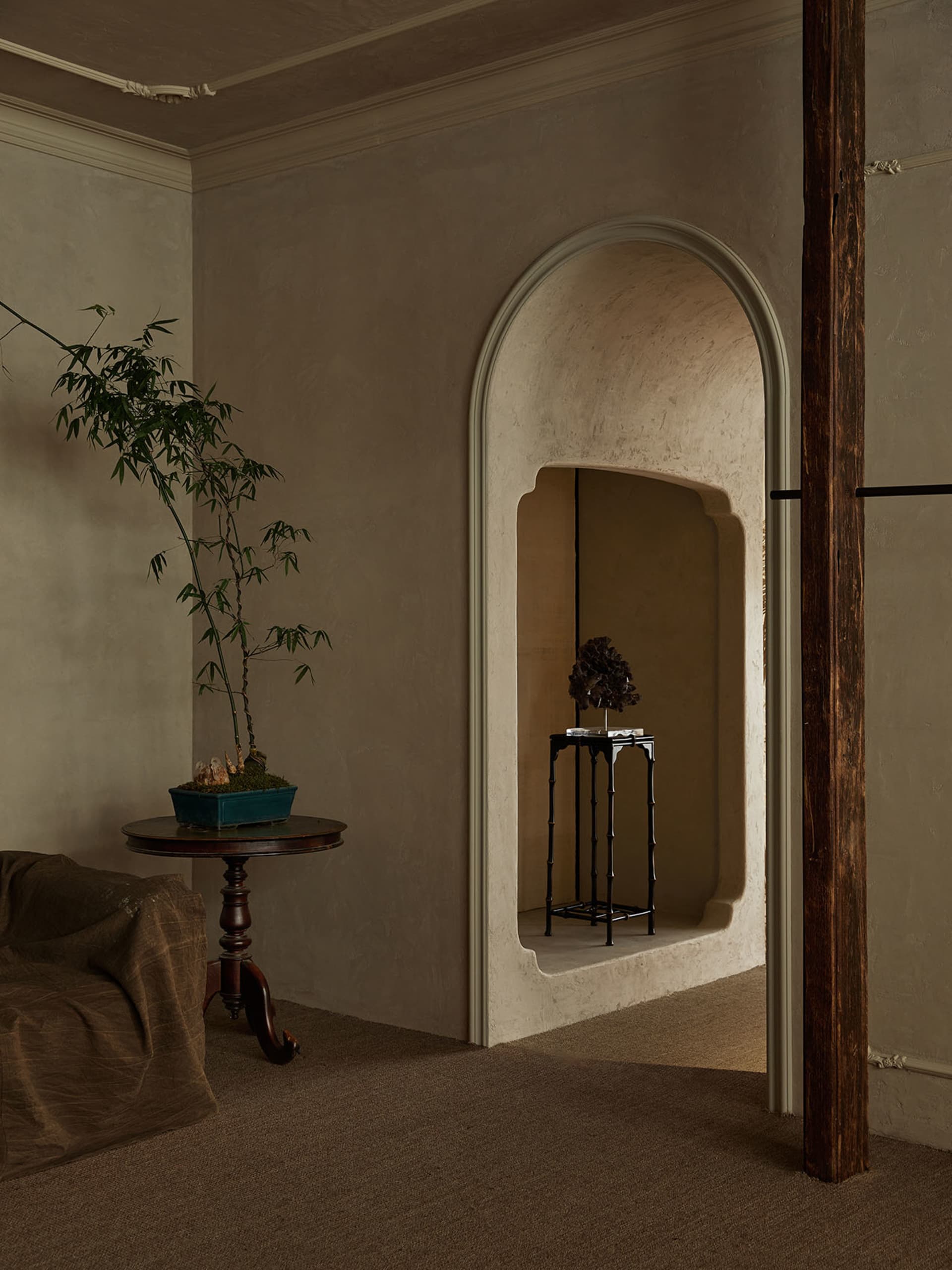 Interior corner of a living room with textured limewash walls, a tall arched decorative niche containing a black metal console and sculptural object, a round wooden side table with a large bamboo plant in a teal planter, and a brown linen sofa in the foreground.